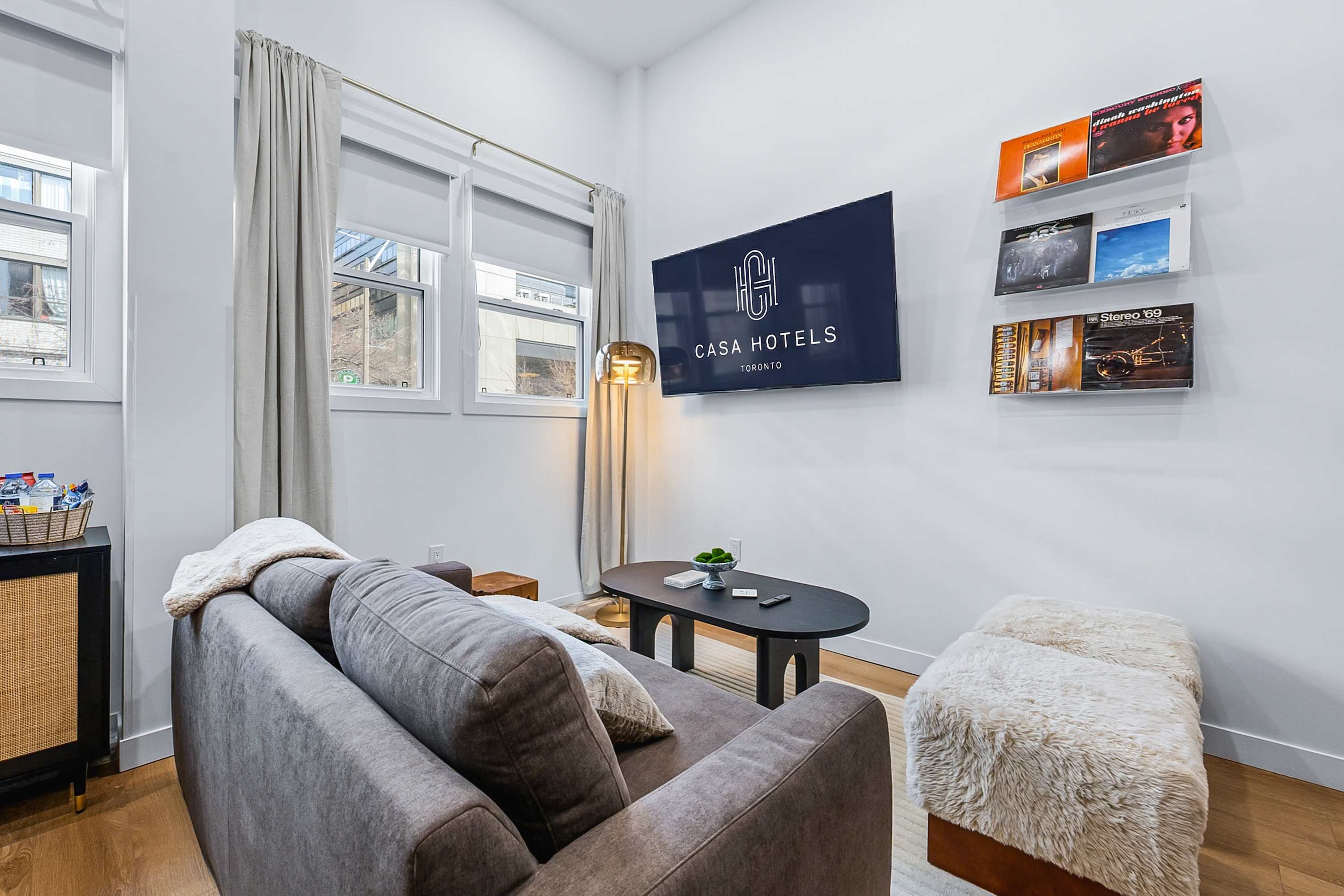Modern living room featuring a grey sofa, a dark oval coffee table, and a large wall-mounted television, complemented by decorative wall shelves displaying records and a stylish gold floor lamp, with natural light streaming through the windows.