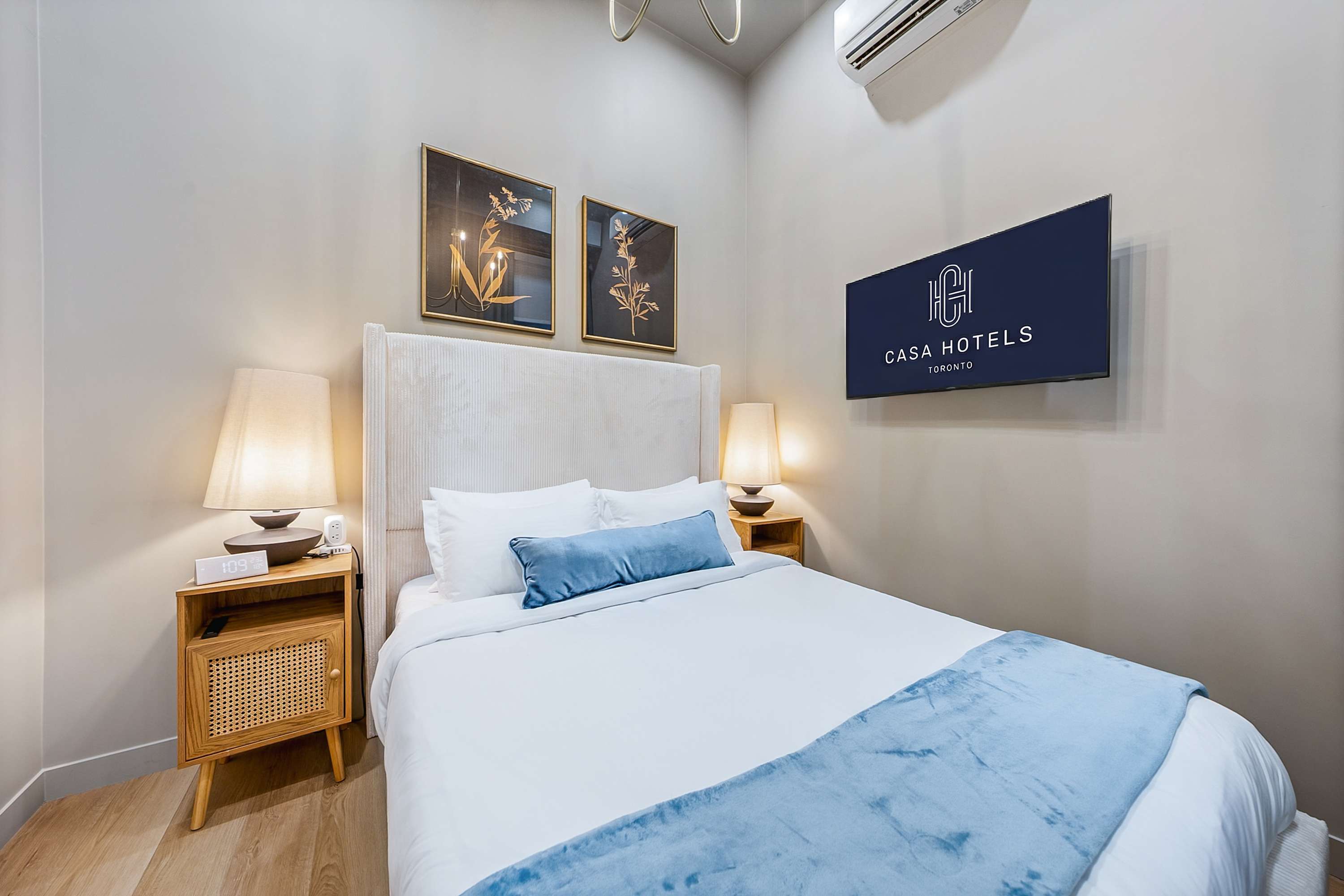 Bright bedroom featuring a queen-sized bed with a tall upholstered headboard, white bedding, and a blue accent pillow, flanked by wooden nightstands with modern lamps, and a wall-mounted TV.
