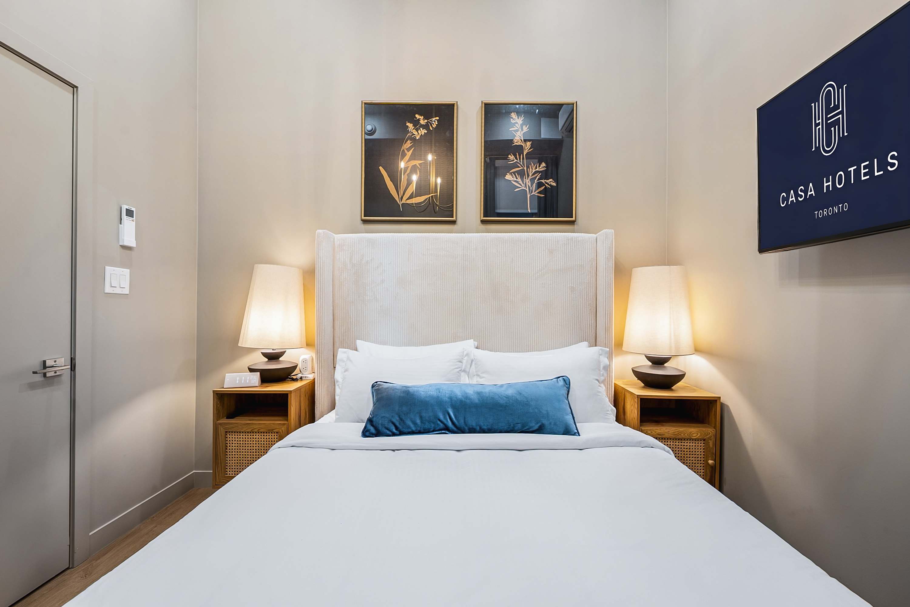 View of the queen-sized bed with a tall upholstered headboard, white bedding, and a blue accent pillow, flanked by wooden nightstands with modern lamps, and two framed artworks above the bed.