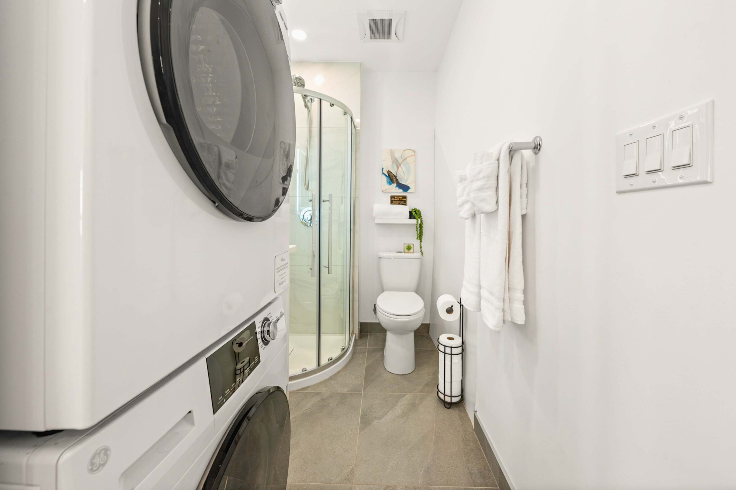 Bathroom view highlighting a stacked white washer and dryer unit, a glass-enclosed corner shower, and a toilet with a small shelf above it, all on grey tiled flooring.