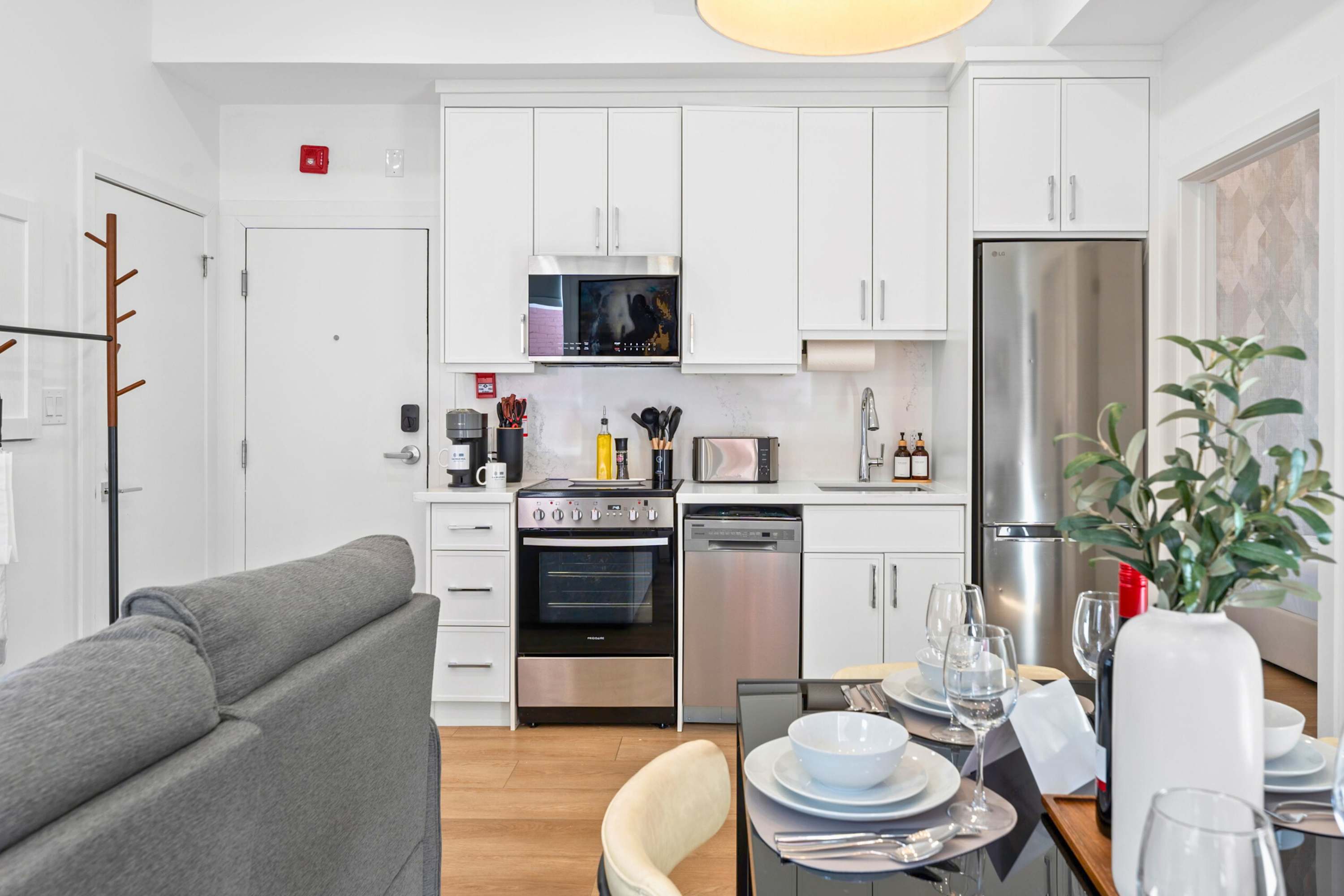 A compact, modern kitchen with white shaker cabinets and stainless steel appliances, including a microwave, stove, and dishwasher, is visible from the living area. A coffee maker, toaster, and various utensils are arranged on the countertop, with a dining table and sofa in the foreground.
