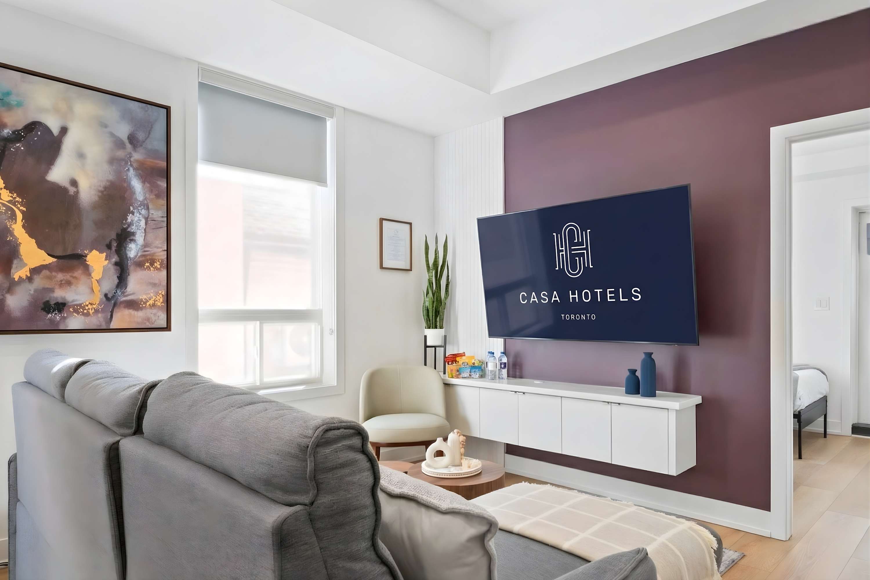 Cozy living room with a grey sectional sofa, a round wooden coffee table, and a cream accent chair. A large TV is mounted on a deep purple accent wall, with a white floating console beneath it and a potted plant nearby.