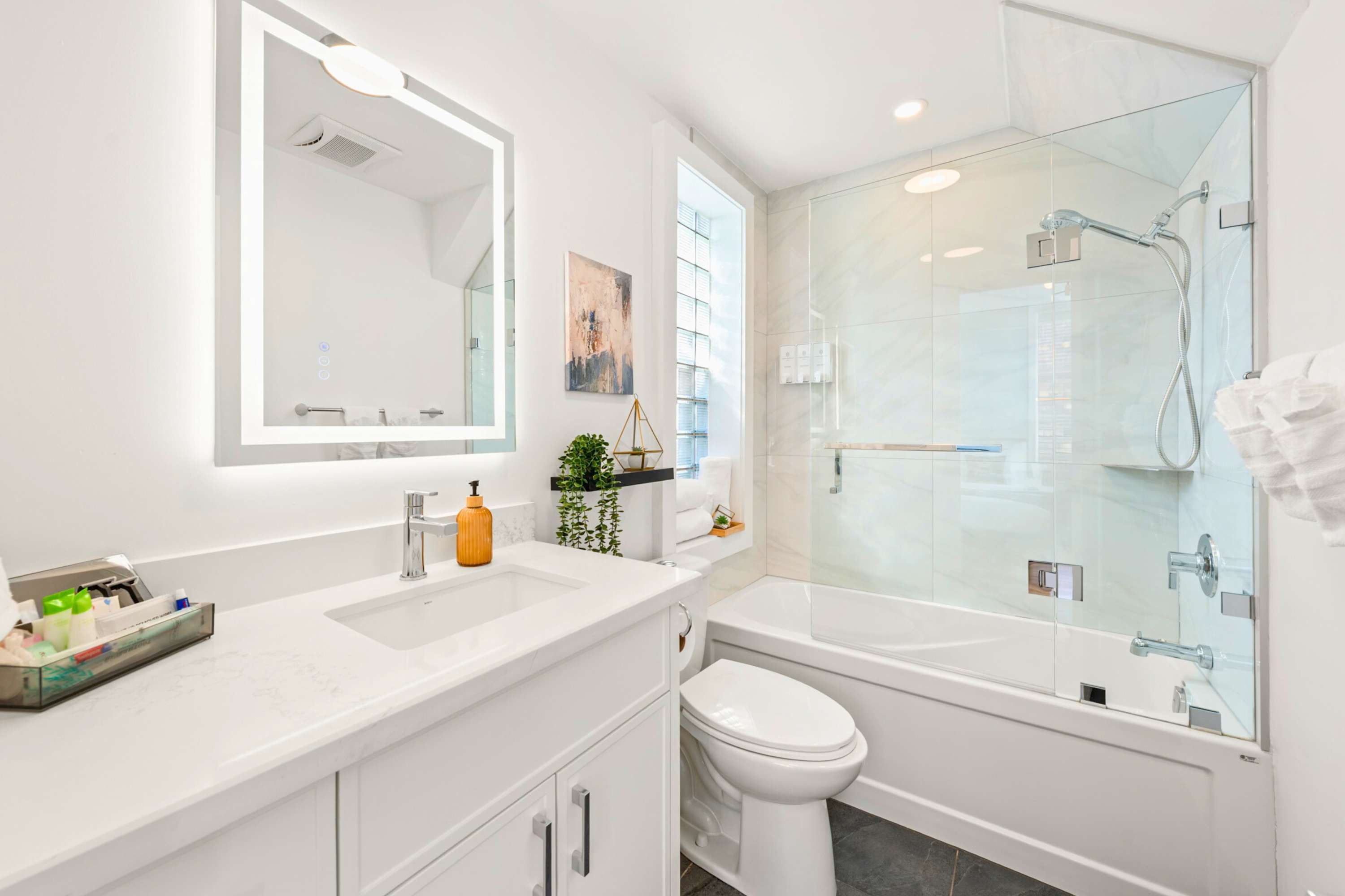 Bright white modern bathroom featuring a single vanity with an illuminated mirror, a toilet, and a glass-enclosed shower/bathtub combo with white tiled walls. A small window with glass blocks is visible.