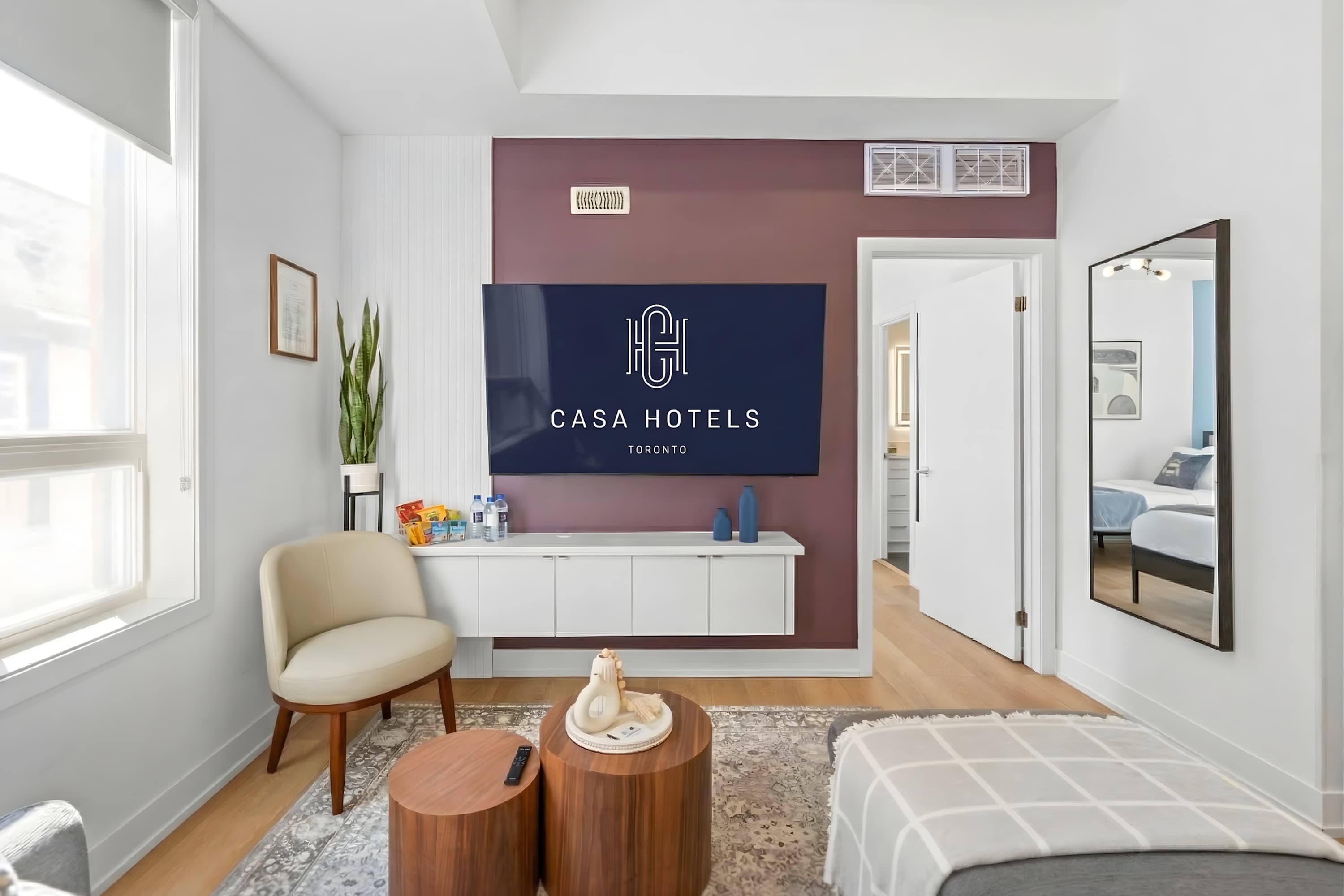 Modern living room featuring a grey sectional sofa, two round wooden coffee tables, and a cream accent chair. A large TV is mounted on a purple accent wall, with a doorway leading to another room visible, reflecting in a full-length mirror.