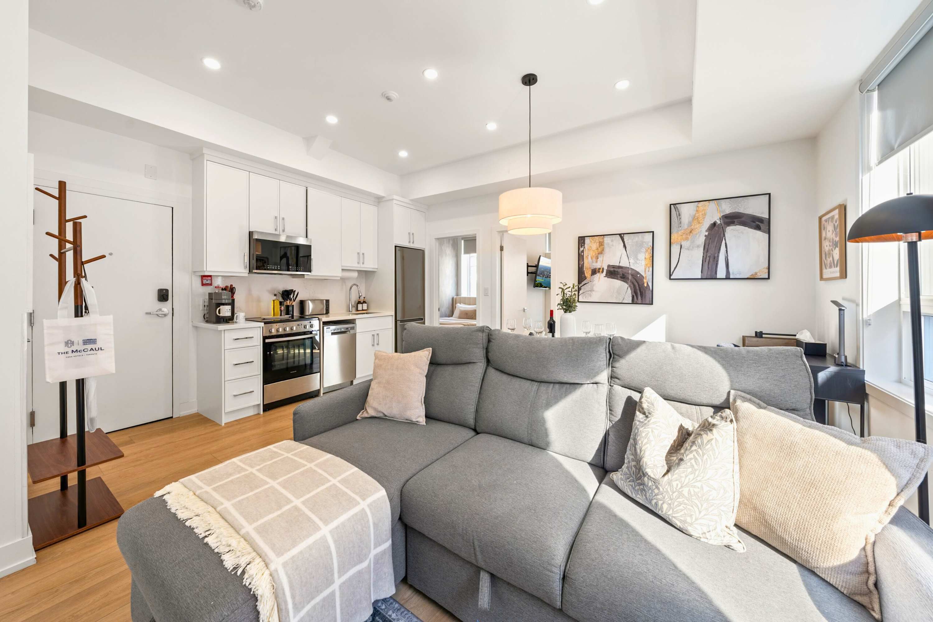 An open-concept living space features a large grey sectional sofa with throw pillows and a blanket, facing a modern kitchen with white cabinetry and stainless steel appliances. A dining area with a black table and light-colored chairs is visible to the right, all under recessed lighting and a contemporary pendant light.