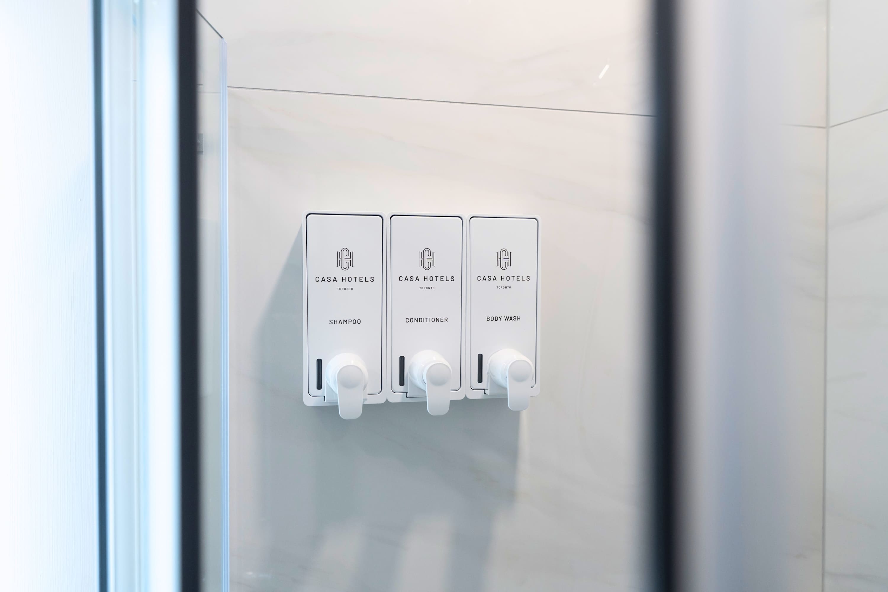Close-up of three white wall-mounted dispensers labeled "SHAMPOO", "CONDITIONER", and "BODY WASH" from "CASA HOTELS Toronto", visible inside a glass shower enclosure.