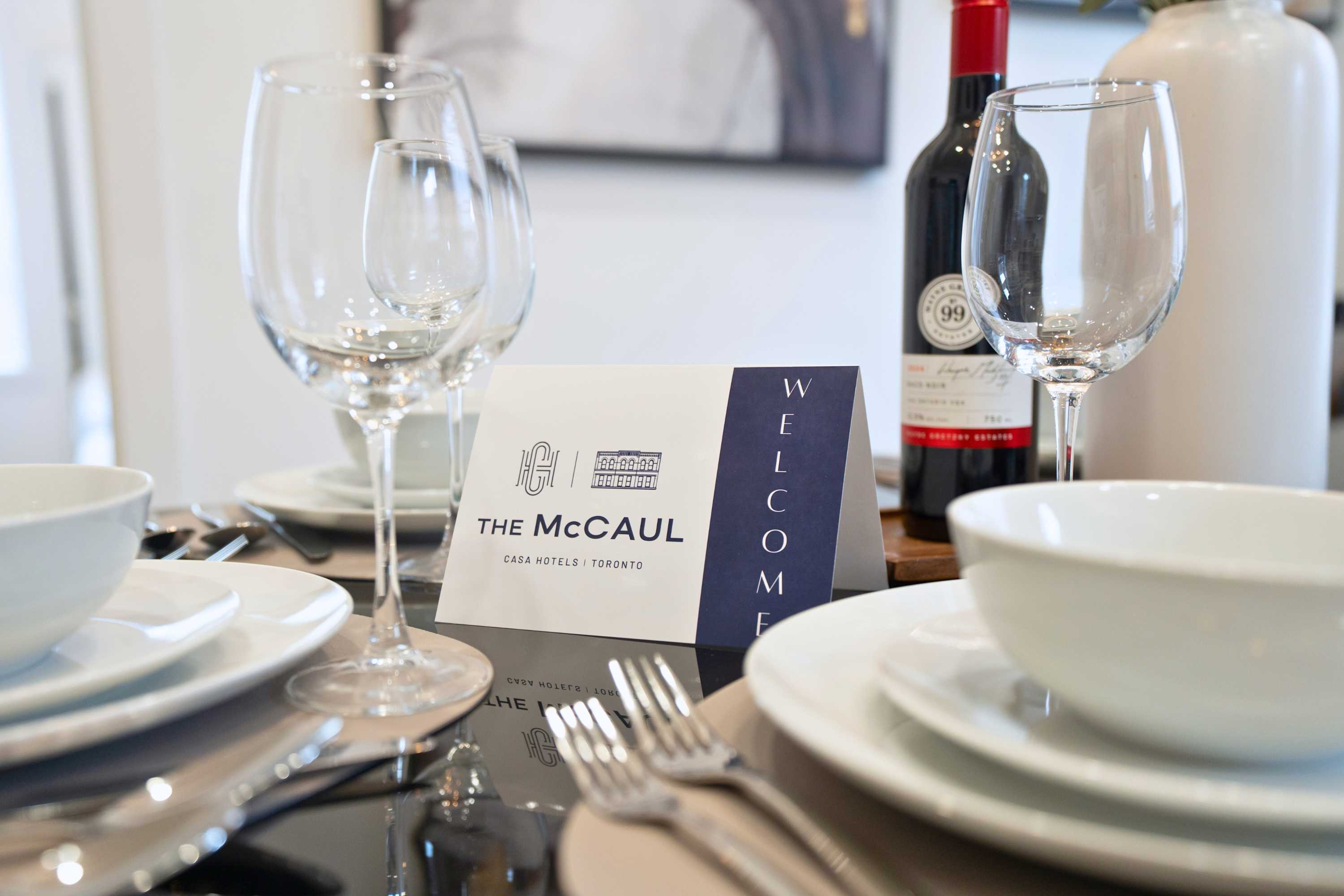 A close-up of a dining table set with white plates, bowls, silverware, and wine glasses, featuring a "WELCOME THE MCCAUL" card and a bottle of red wine. A small green plant in a white vase adds a touch of nature to the elegant setting.