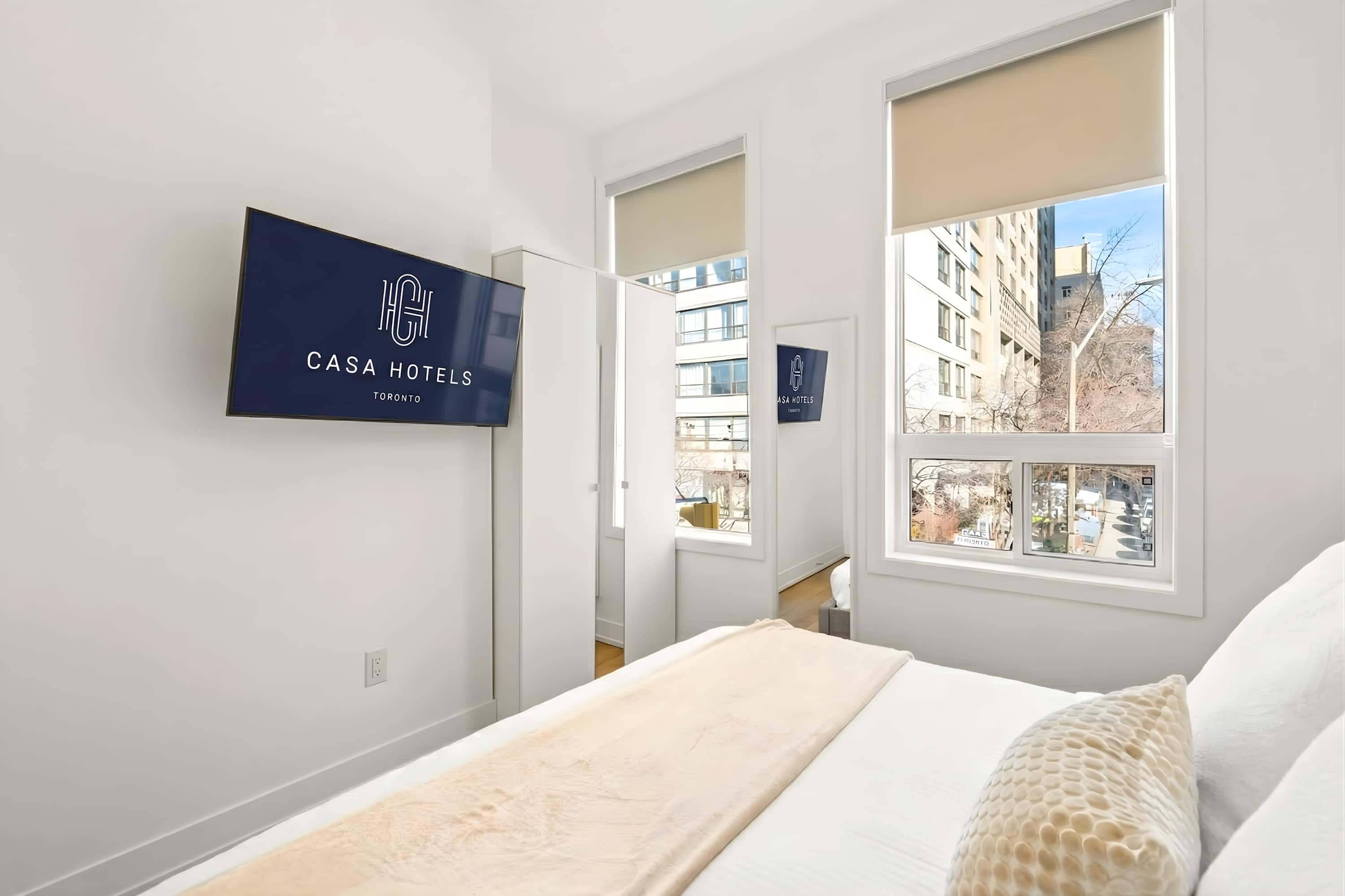 Bright bedroom featuring a wall-mounted TV displaying 'CASA HOTELS Toronto', a white bed with light bedding, and two large windows with roller shades offering a city view.