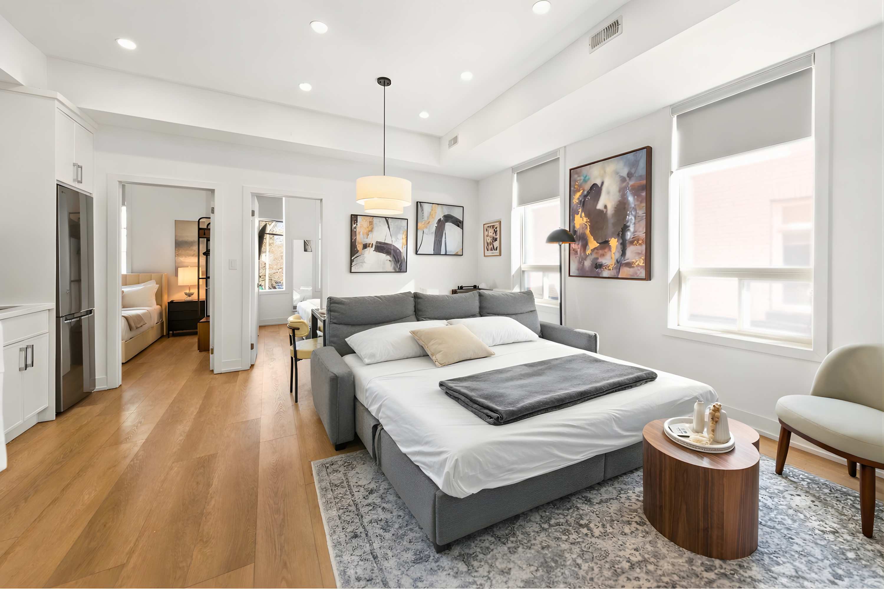 The living room features a grey sectional sofa converted into a comfortable pull-out bed, positioned on a patterned area rug. Light hardwood floors and large windows frame the space, with abstract artwork on the walls.