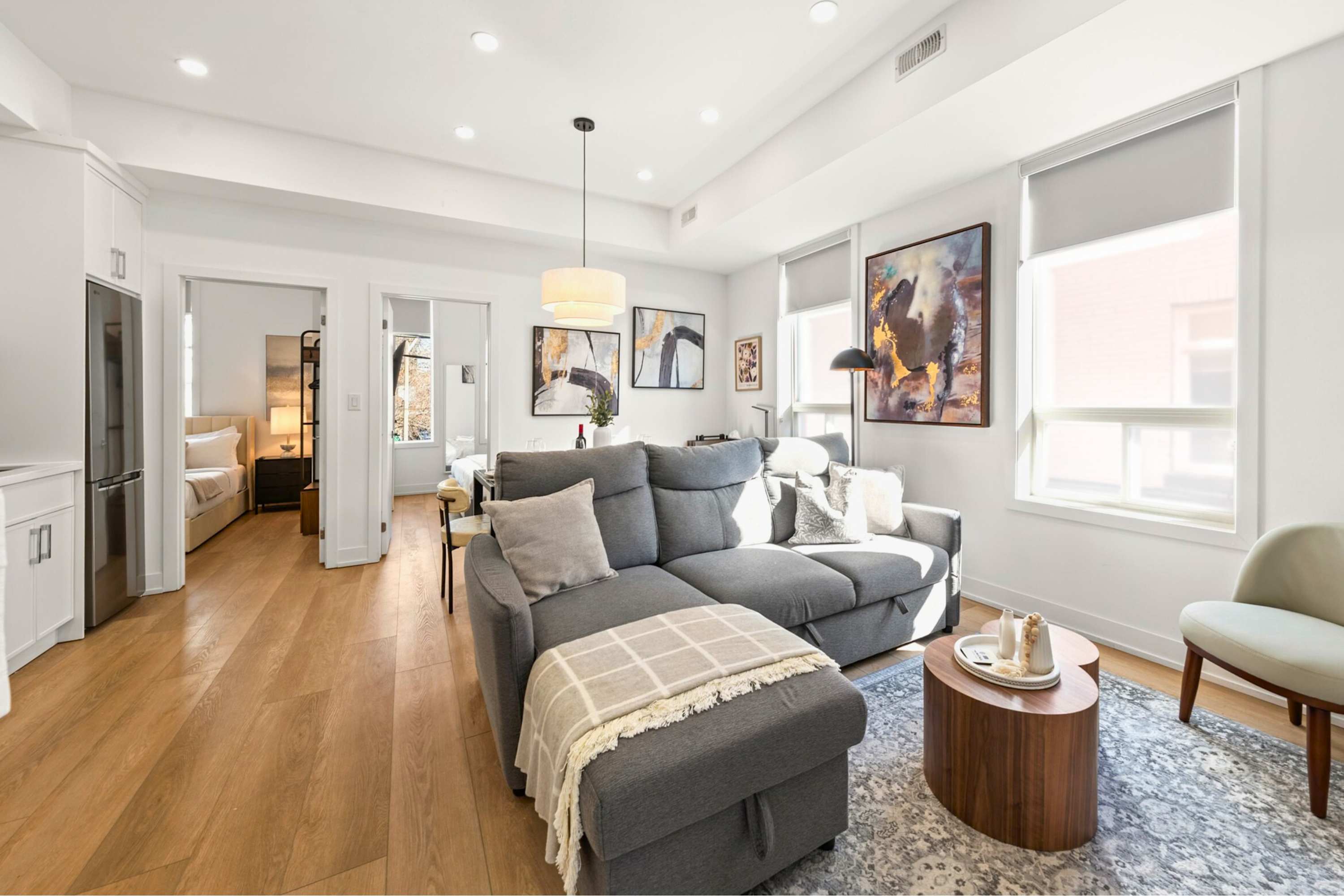 Spacious open-concept living and dining area with a grey sectional sofa, a black dining table, and light hardwood floors. Large windows provide natural light, and abstract artwork adorns the walls, creating a modern aesthetic.