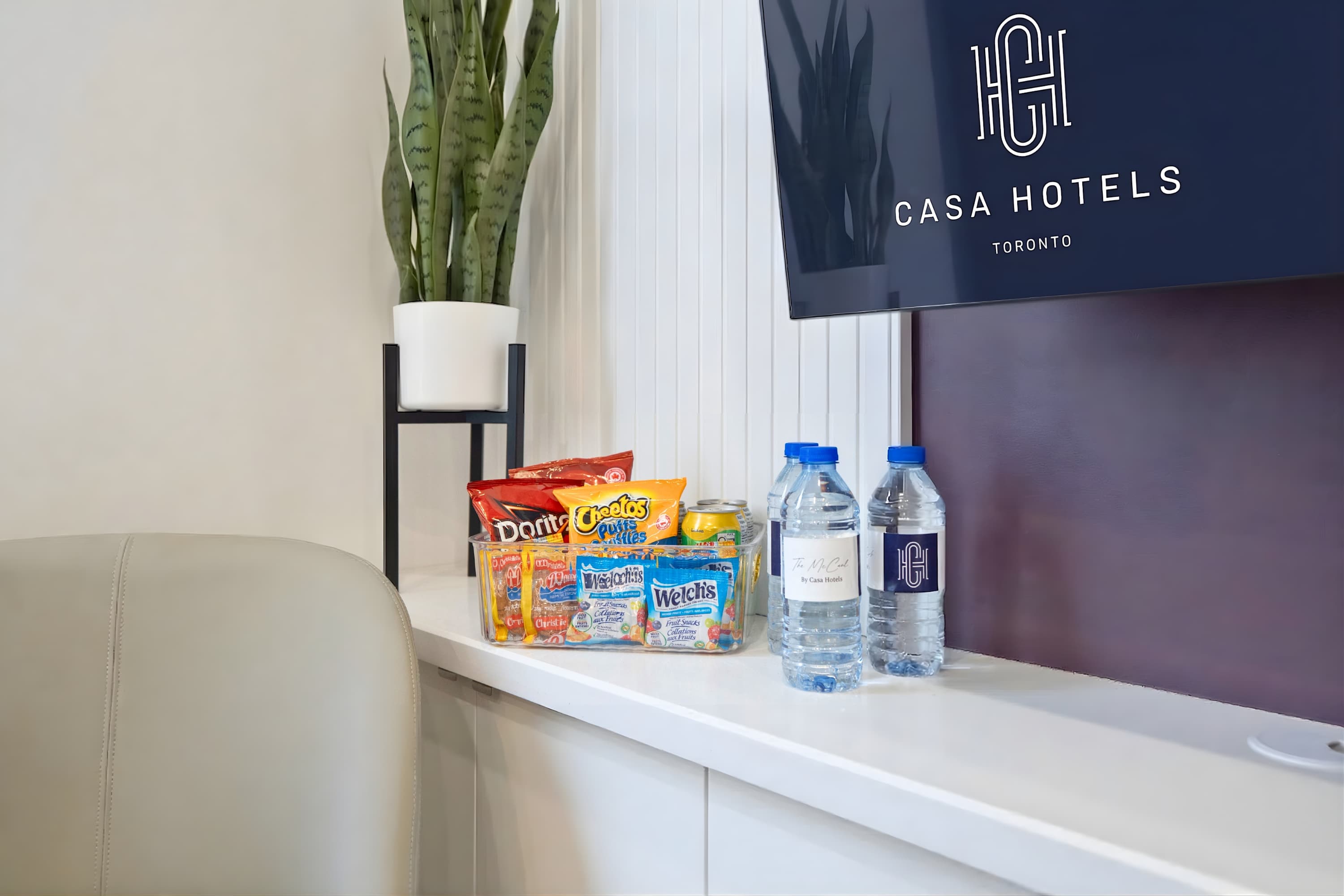 A clear plastic bin filled with various snack bags and two bottled waters sits on a white shelf below a wall-mounted TV displaying "CASA HOTELS TORONTO" logo. A tall snake plant in a white pot on a black stand adds greenery to the corner.