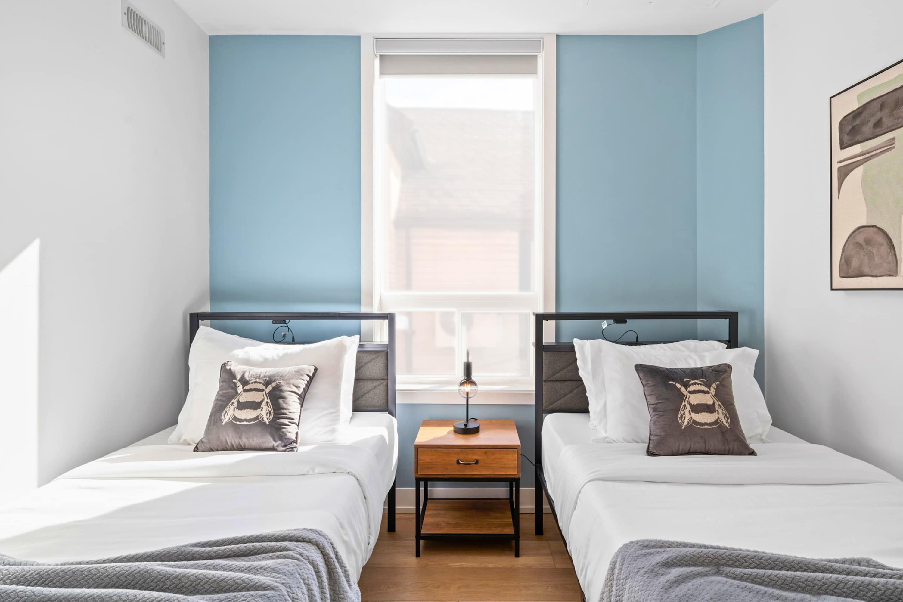 Close-up view of the bedroom with two single beds, each featuring a dark grey pillow with a bee motif, a shared wooden nightstand, and a modern table lamp.