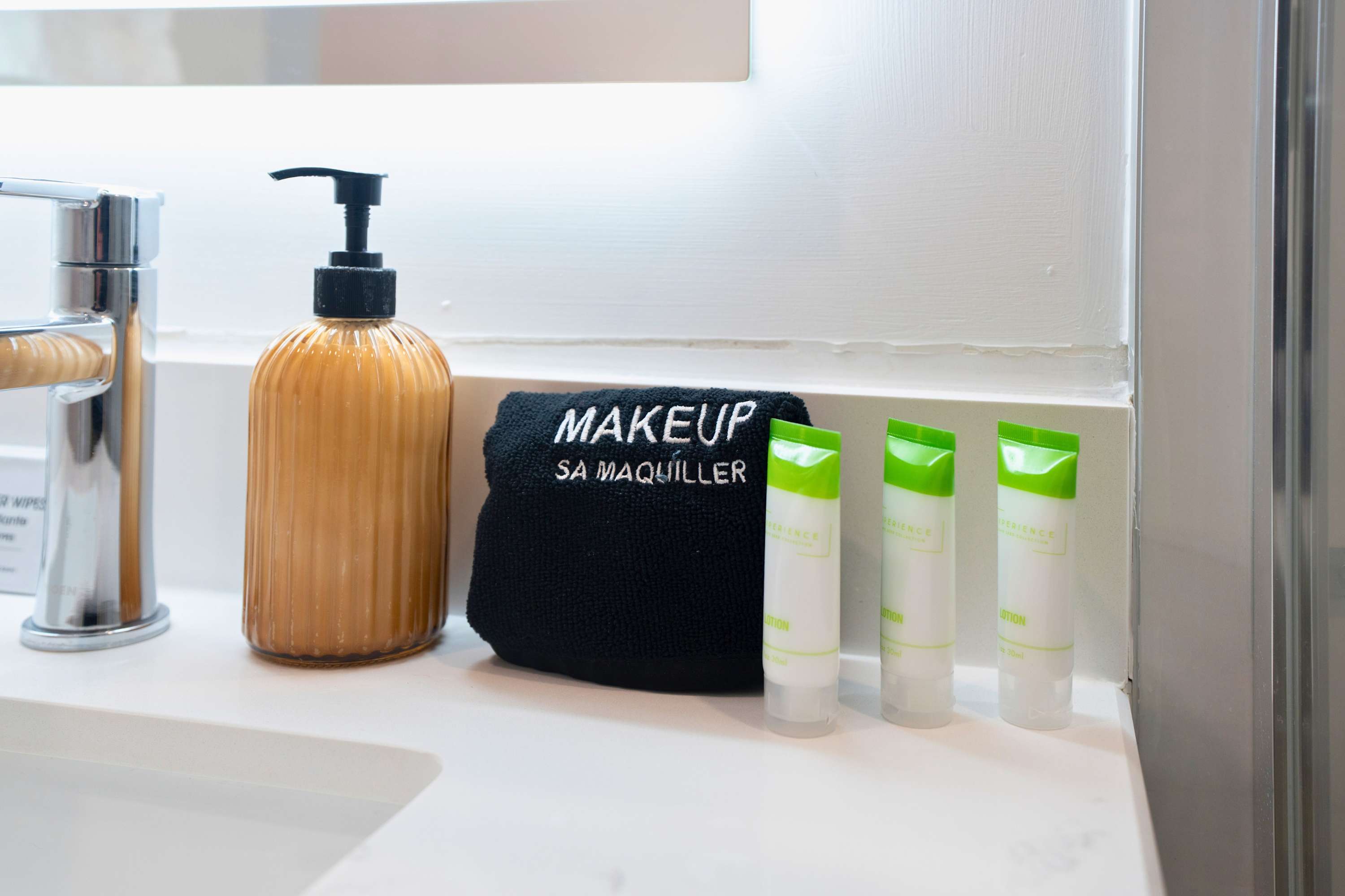 Detailed view of the bathroom countertop showcasing a ribbed amber soap dispenser, a black 'MAKEUP' towel, and three small green-capped lotion bottles next to a modern faucet.
