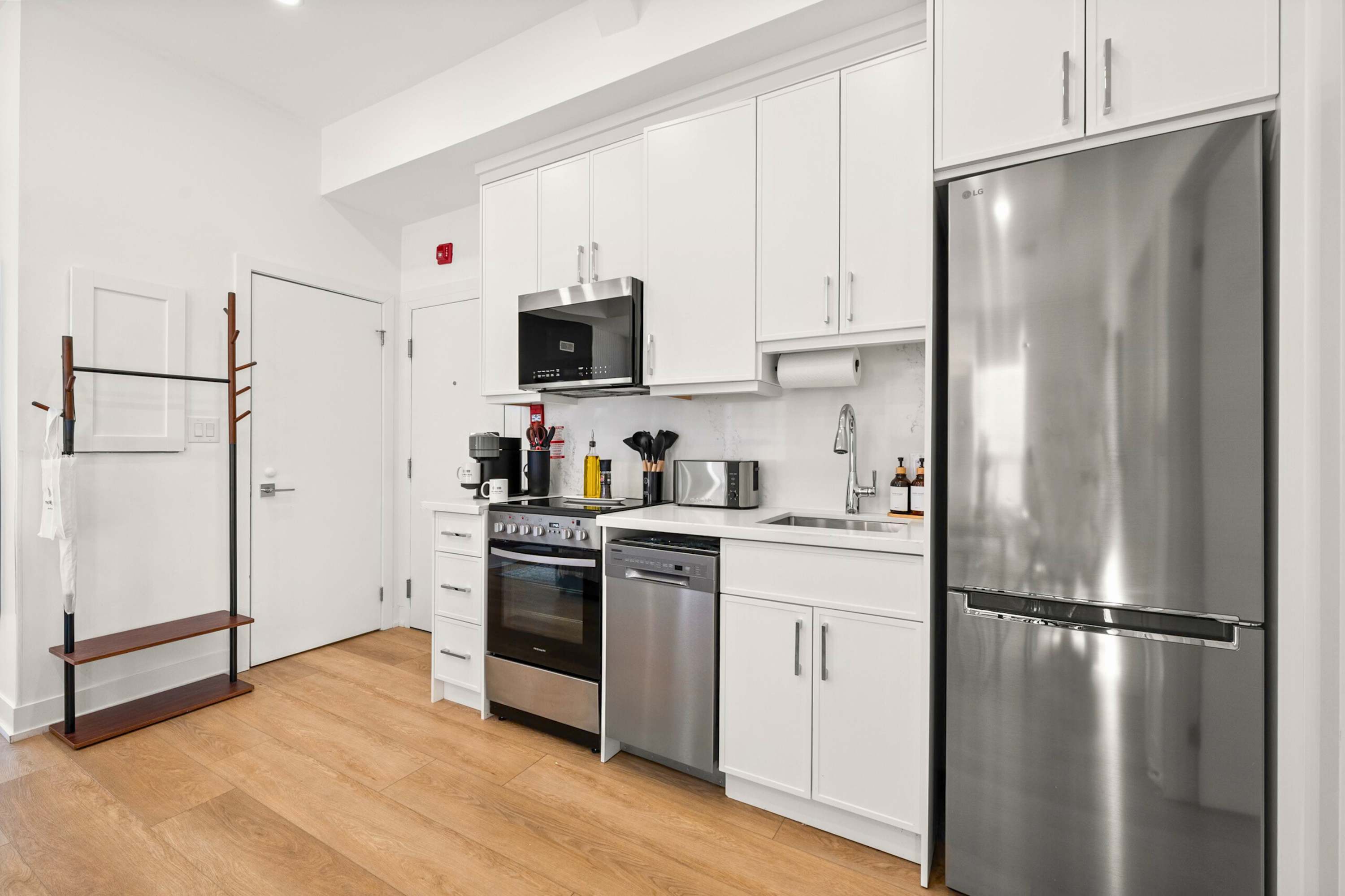 A well-equipped modern kitchen features white upper and lower cabinets, stainless steel appliances including a refrigerator, microwave, stove, and dishwasher, and light wood flooring. A coffee maker and toaster are visible on the countertop, with a paper towel holder above the sink.