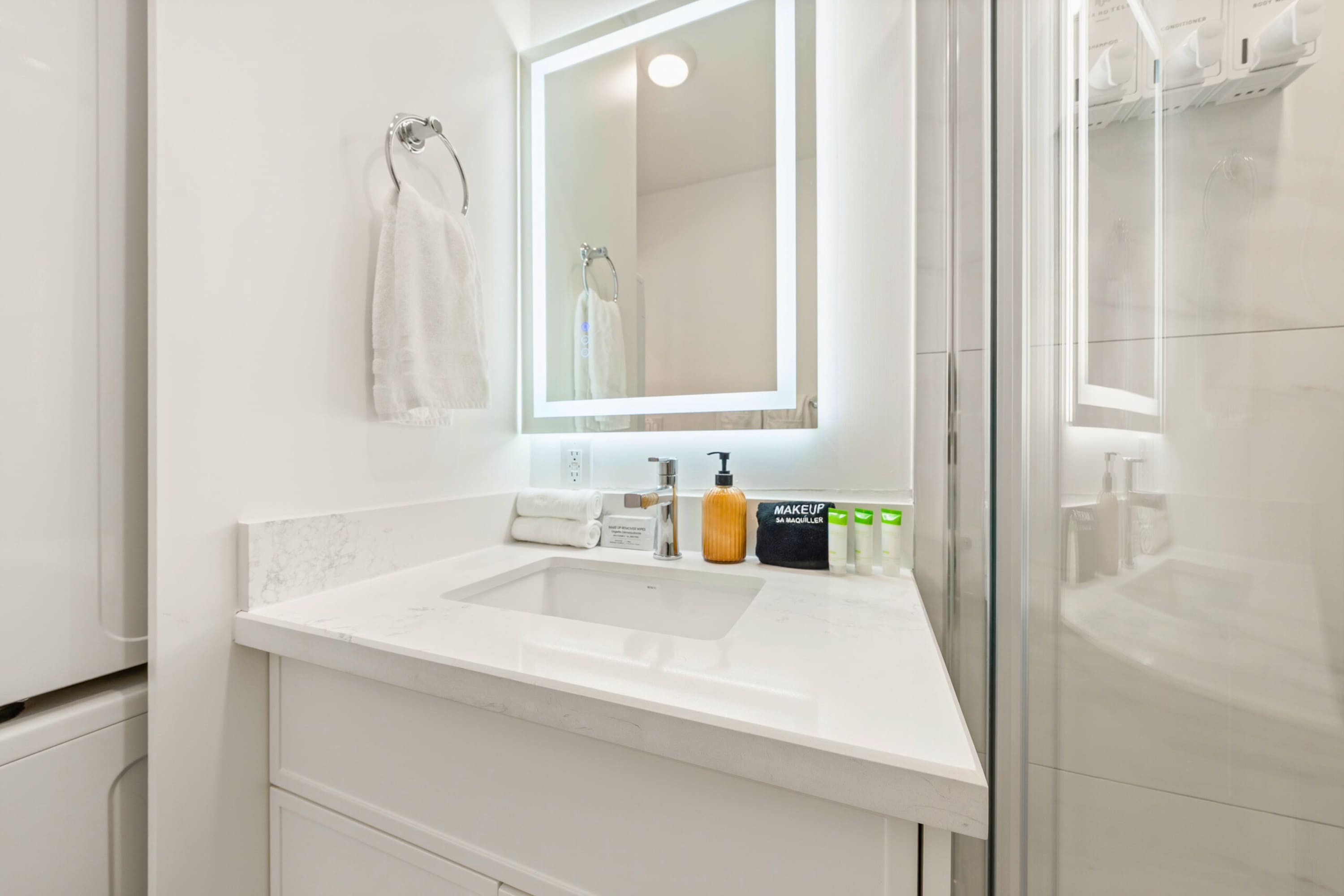 Close-up of the bathroom vanity featuring a white countertop, integrated sink, modern faucet, illuminated mirror, and various toiletries including a soap dispenser and lotions.