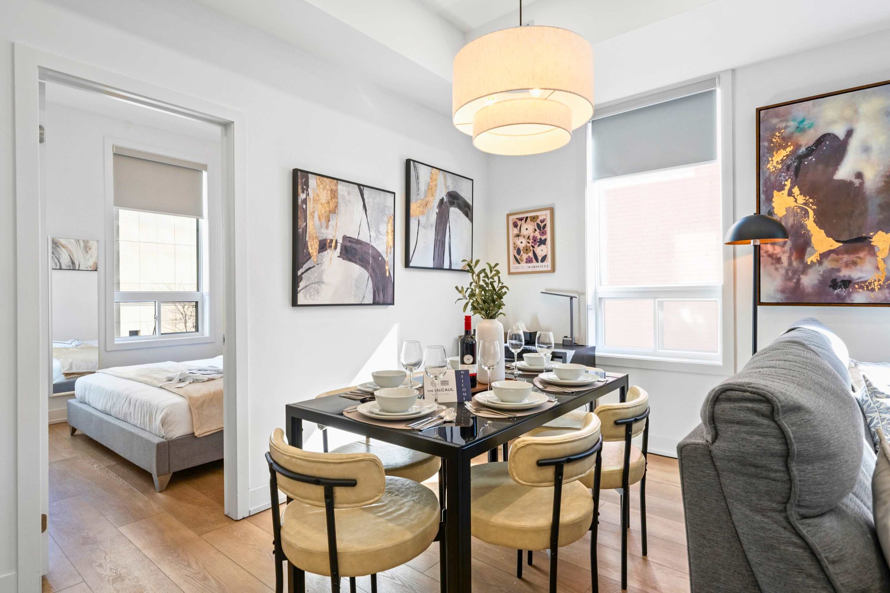 A modern dining area features a black rectangular table set for four with light-colored chairs, illuminated by a multi-tiered pendant light. Abstract art adorns the wall, and a bedroom is visible through an open doorway, with large windows providing natural light.