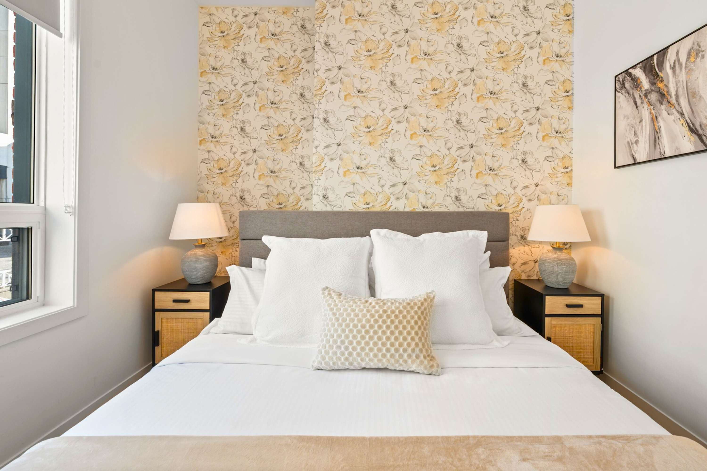 A cozy bedroom features a bed with white linens and a textured throw, flanked by two modern nightstands with woven fronts and grey ceramic lamps. A striking yellow floral wallpaper accent wall serves as a headboard, complemented by an abstract art piece.