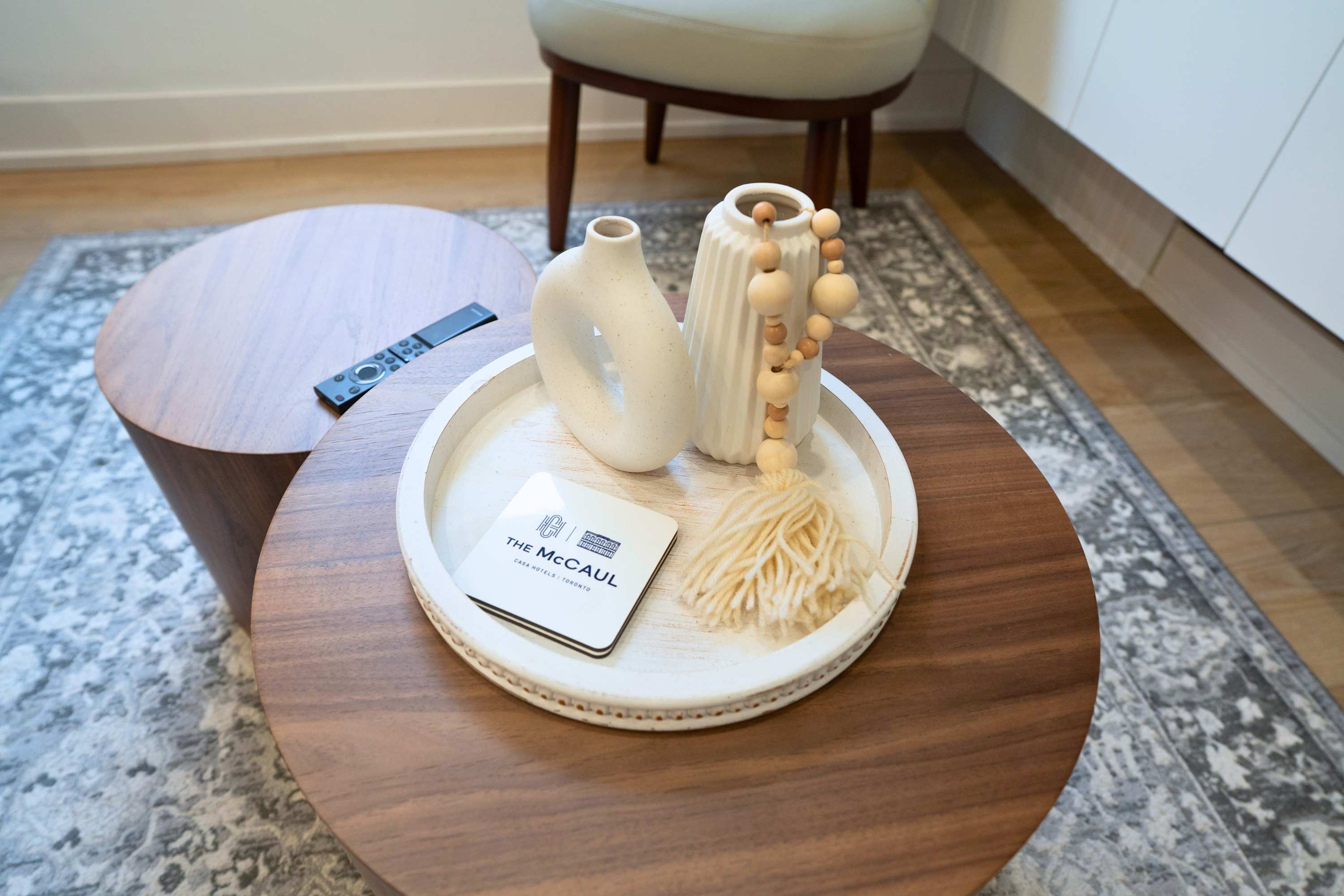 A close-up of two round wooden coffee tables, one light and one dark, featuring a white decorative tray with two ceramic vases, wooden beads, and coasters. A remote control rests on the lighter table, all set on a patterned grey rug.