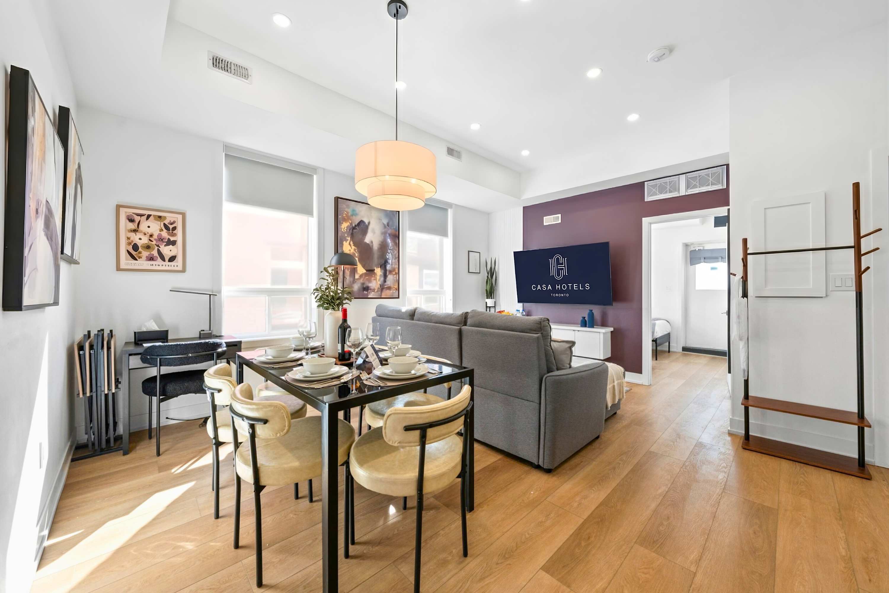 Bright open-concept living and dining area featuring a black dining table set for four, a grey sectional sofa, and a wall-mounted TV against a deep purple accent wall. A small desk with a lamp is visible in the corner.