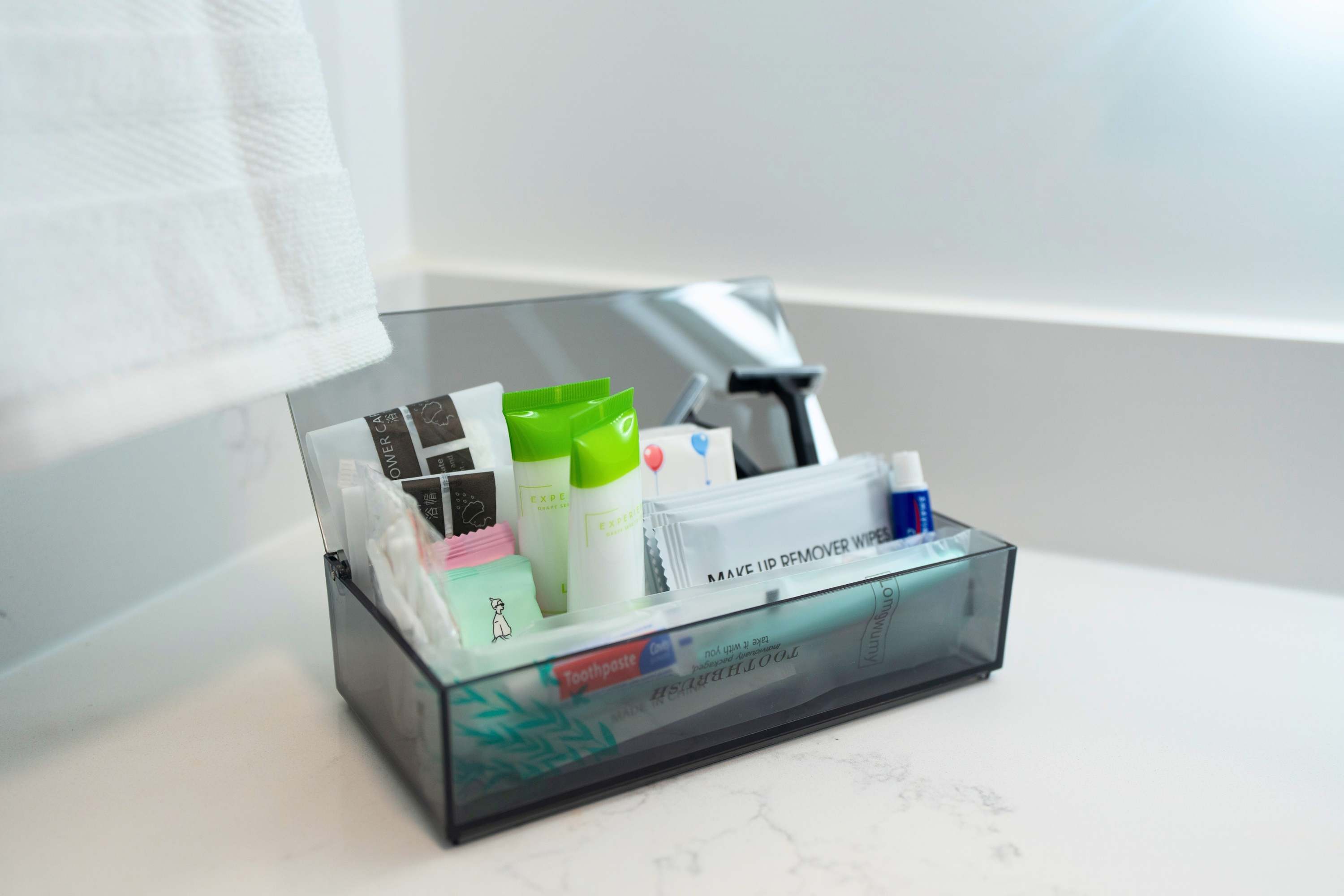 Close-up view of a clear acrylic organizer filled with various travel-sized toiletries, including toothpaste, razors, makeup remover wipes, and cotton swabs, resting on a white countertop.