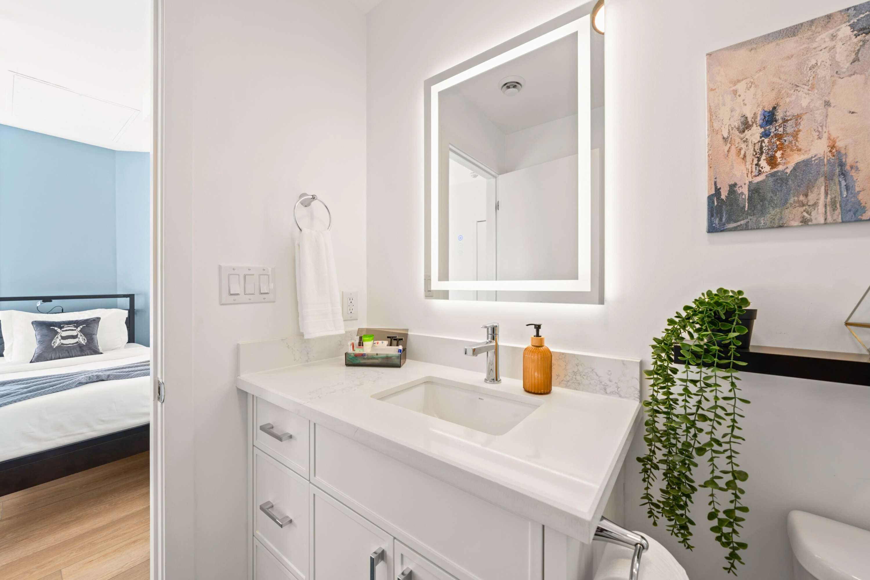Modern white bathroom vanity with a rectangular illuminated mirror, a white quartz countertop, and a chrome faucet. A glimpse of a blue-walled bedroom with a bed is visible through the open doorway.