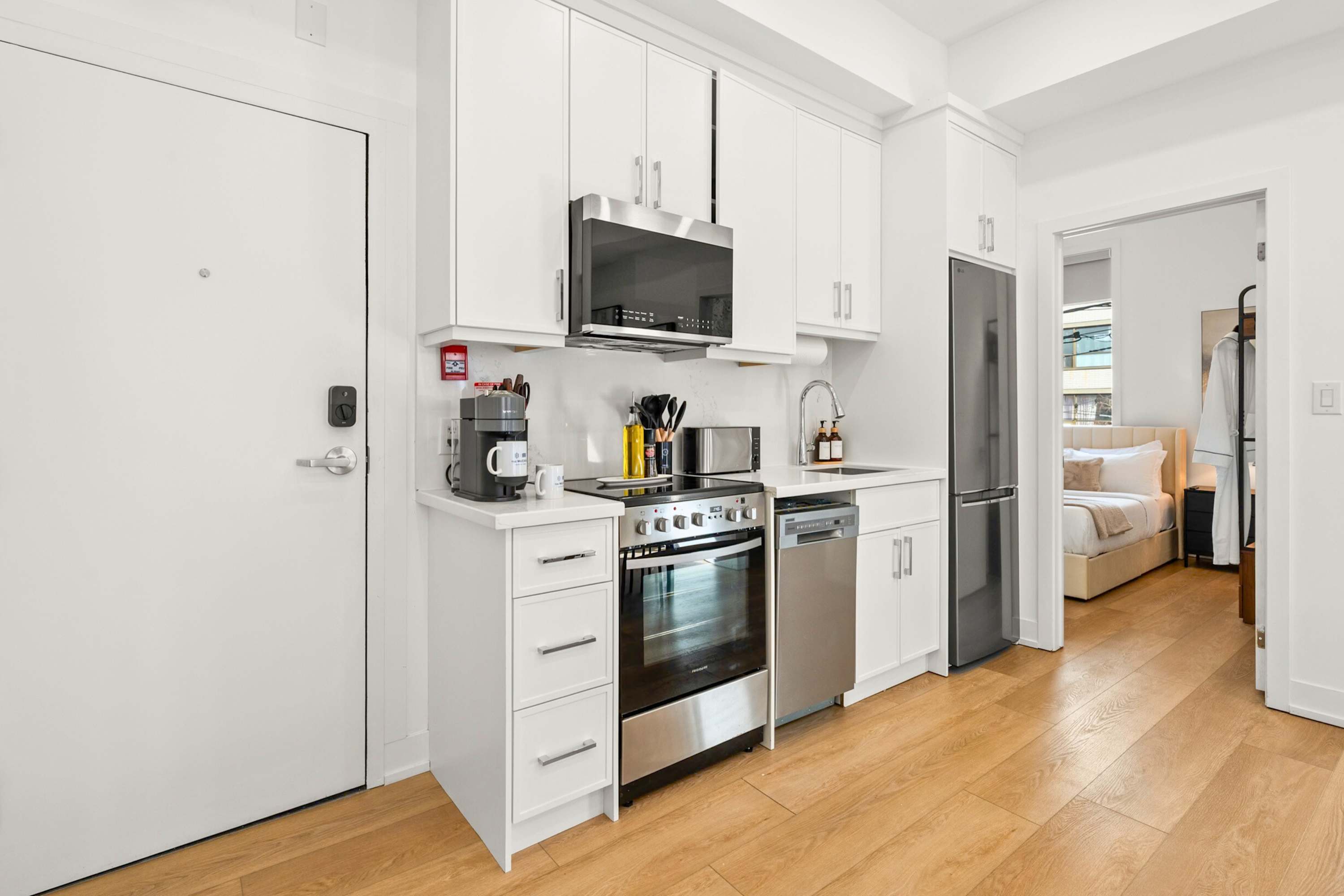 A bright, modern kitchen with white cabinetry and stainless steel appliances, including a refrigerator, stove, and microwave, is shown from an angle that reveals the entrance door with a keyless lock. Hardwood floors extend throughout the space, with a glimpse into a bedroom beyond.