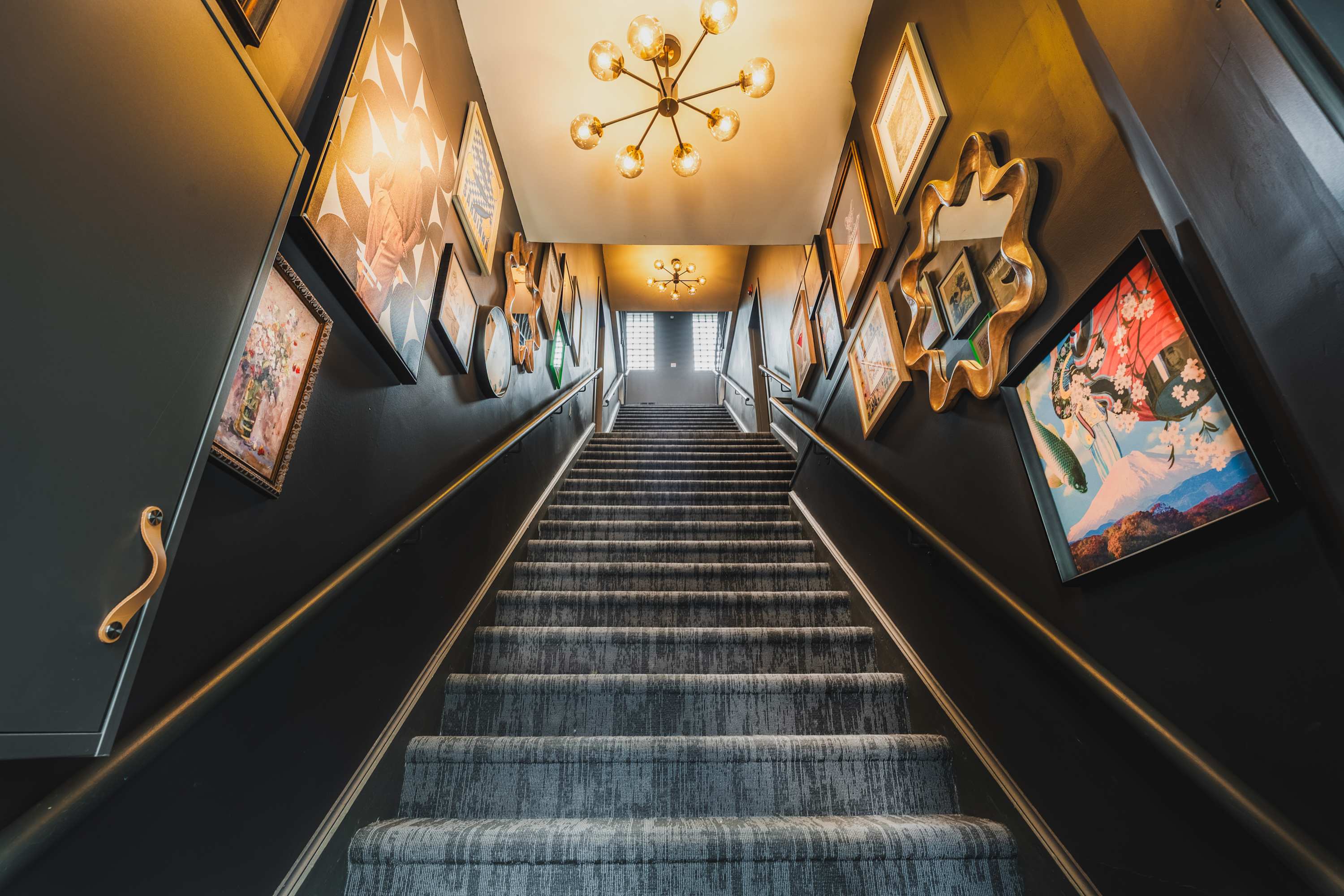 View looking up a wide carpeted staircase with dark grey walls decorated with an eclectic collection of framed art and mirrors. Multiple modern gold chandeliers provide lighting.