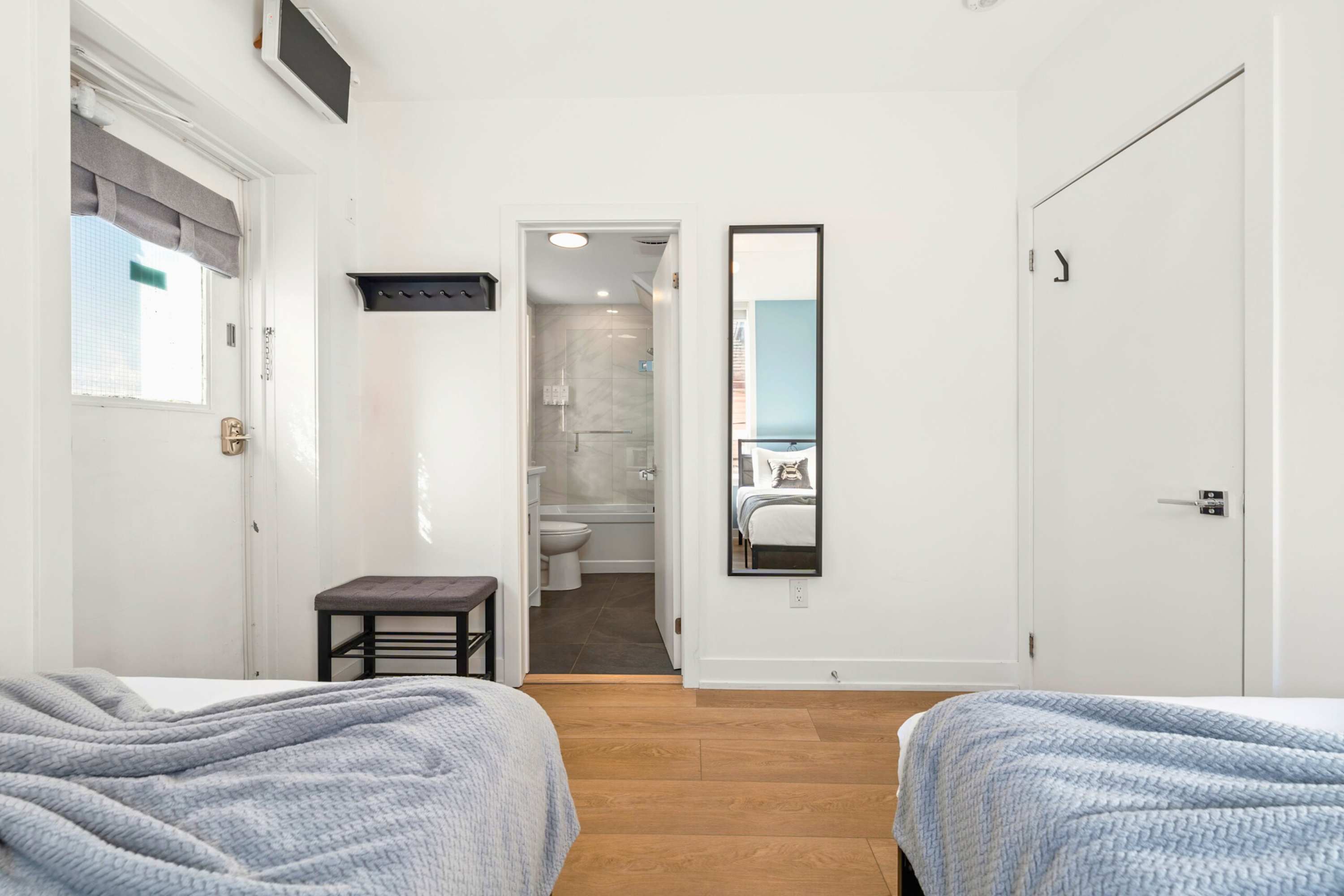 View from the bedroom with two single beds, looking towards a doorway that leads to a bathroom, featuring light hardwood floors and a full-length mirror on the wall.