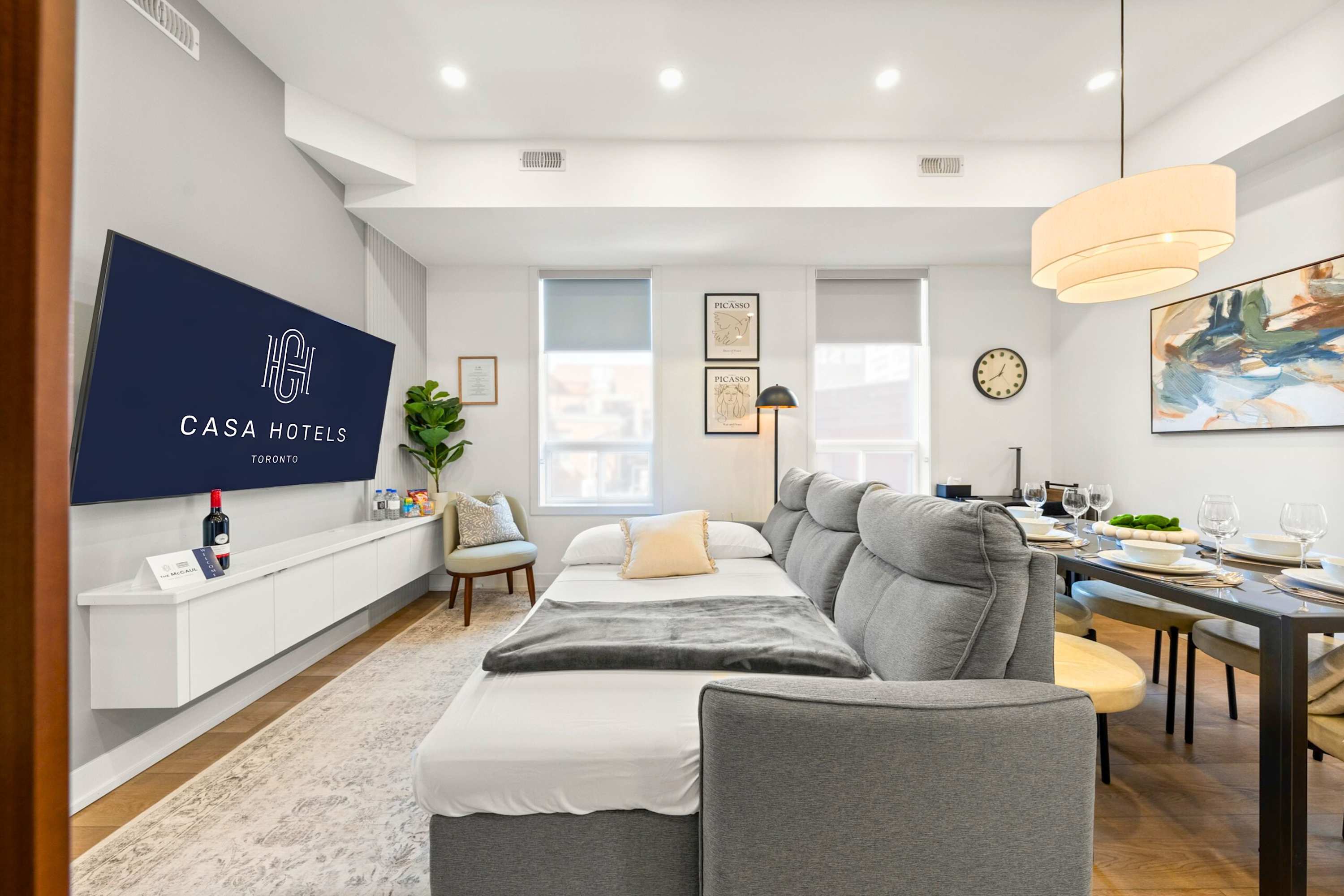 Bright living room featuring a large grey sectional sofa that converts into a bed, a wall-mounted TV displaying "CASA HOTELS", and a white floating media console. Two large windows with roller shades provide natural light, and a dining area is visible in the background.