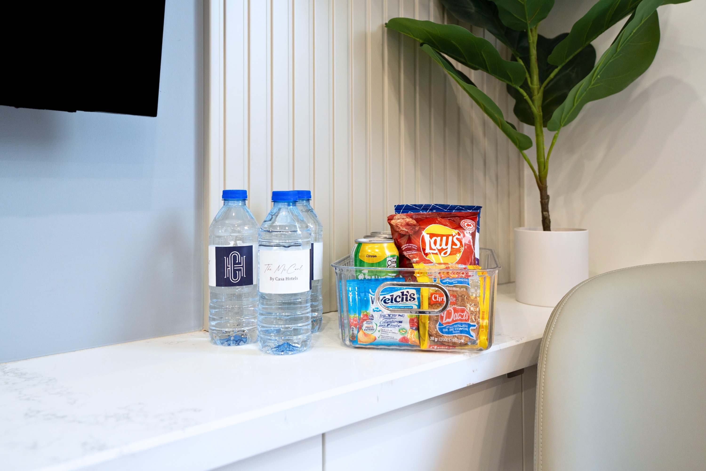 Close-up of a white floating console in the living room, displaying two bottled waters with "CASA HOTELS" labels and a clear container filled with various snacks like chips, candy, and juice boxes. A small indoor plant is visible in the background.