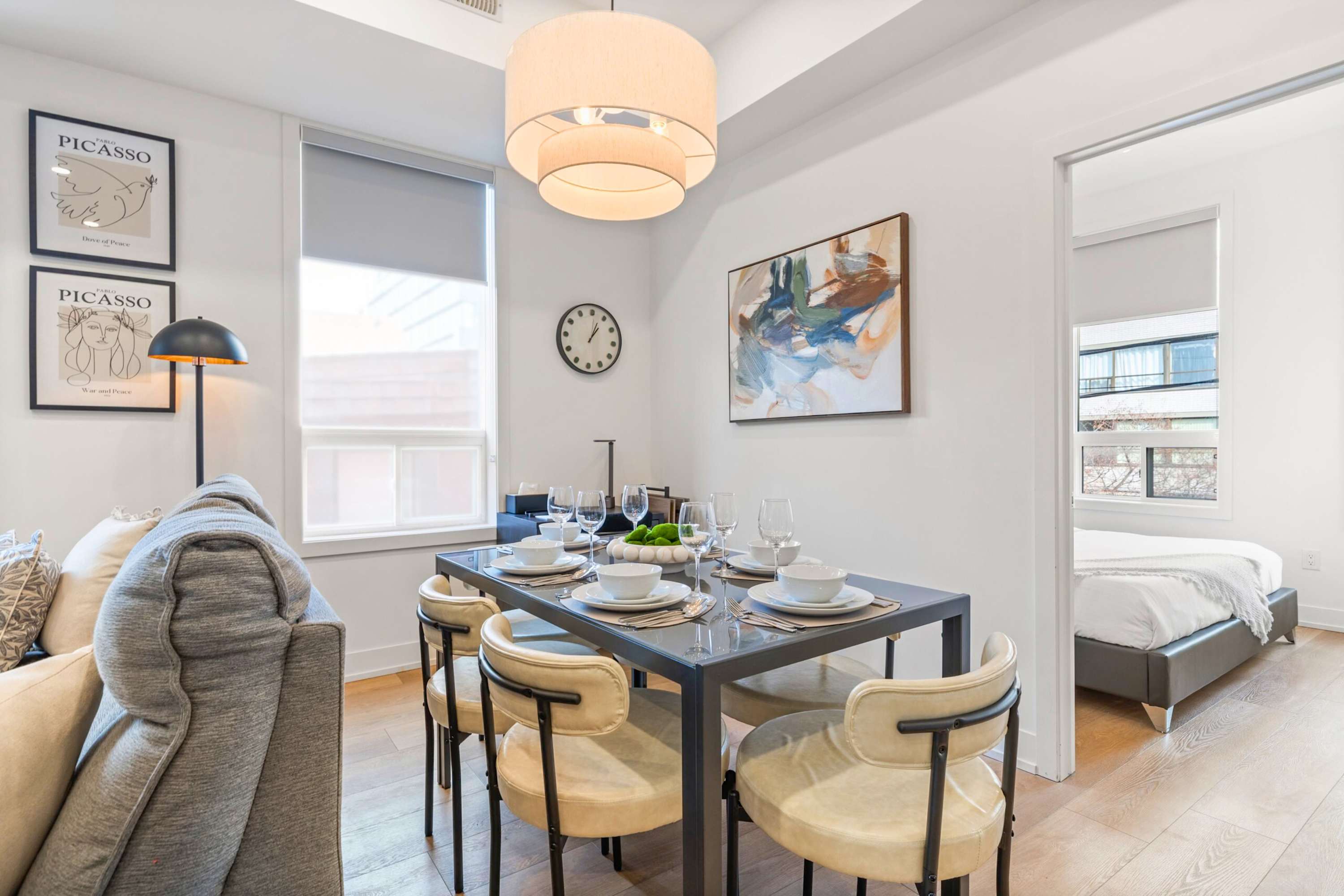Modern dining area with a dark rectangular table set for four, featuring white place settings and light-colored upholstered chairs. A large drum pendant light hangs above, and abstract artwork adorns the wall.