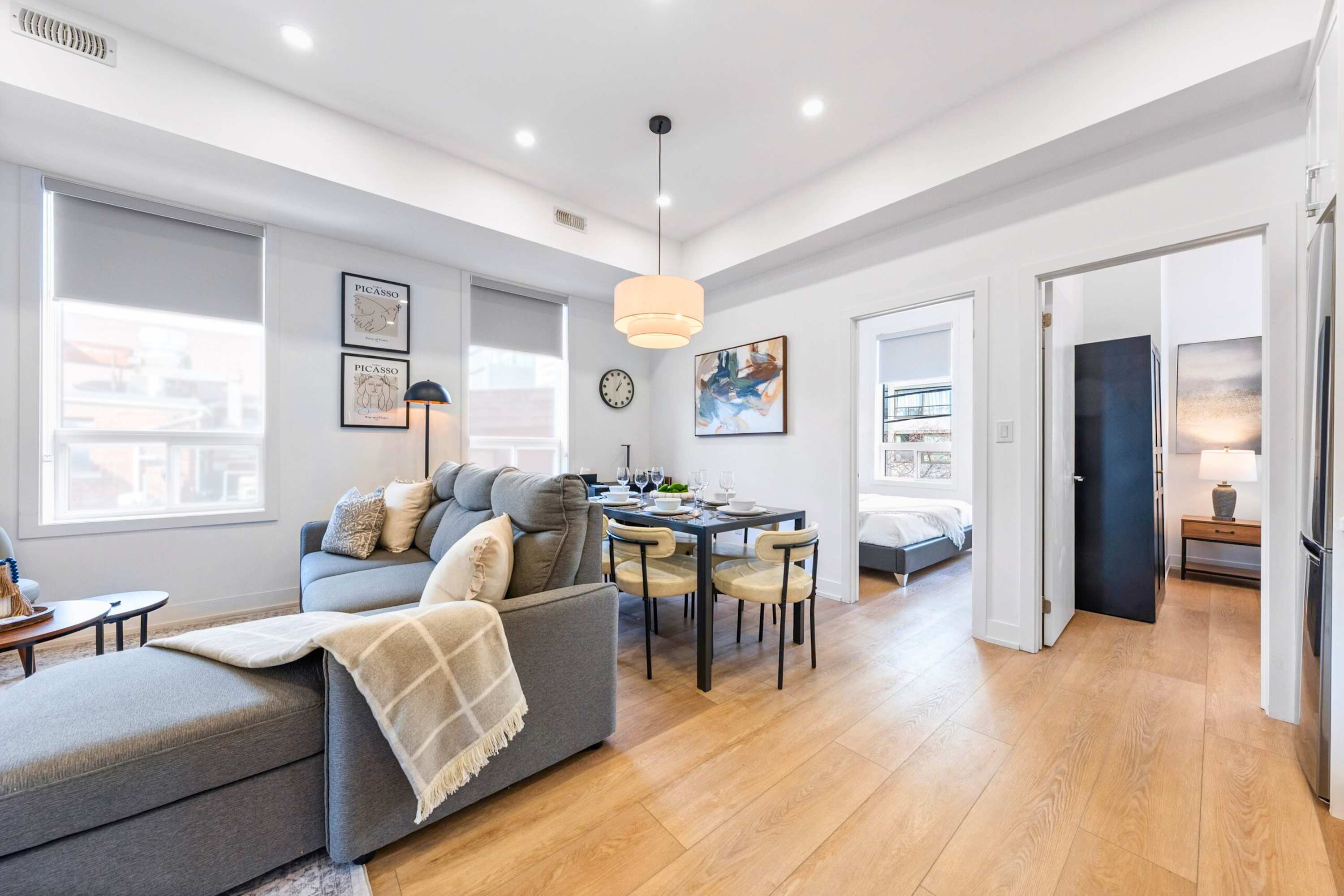 Expansive open-concept living space with a grey sectional sofa, a dining area, and light hardwood floors. Large windows with roller shades illuminate the room, and a doorway leads to a bedroom in the background.