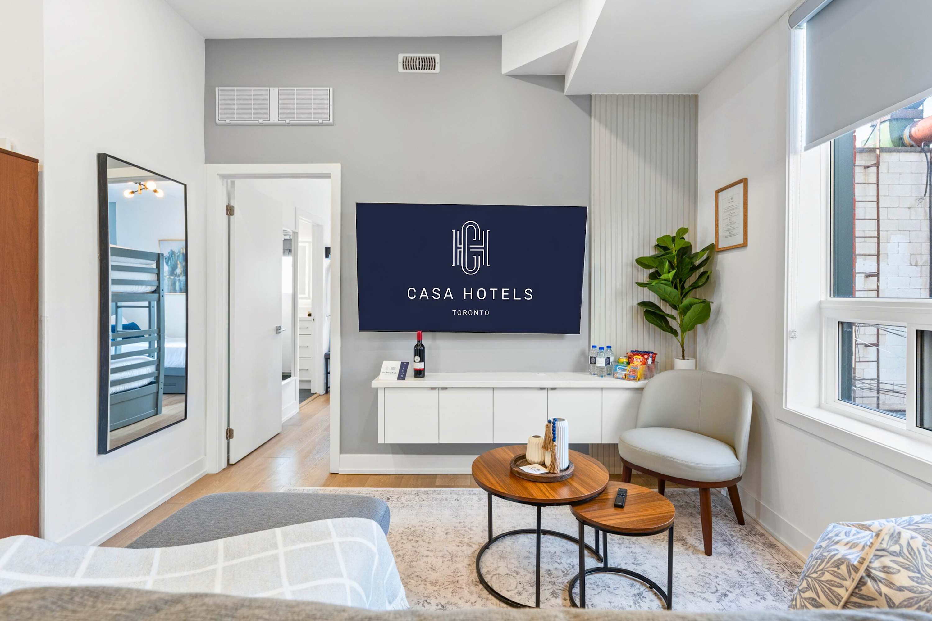 Cozy living room featuring a grey sectional sofa, a light-colored accent chair, and two round nesting coffee tables on a patterned rug. A large wall-mounted TV with "CASA HOTELS" branding is centered above a white floating console, with a plant and welcome amenities nearby.