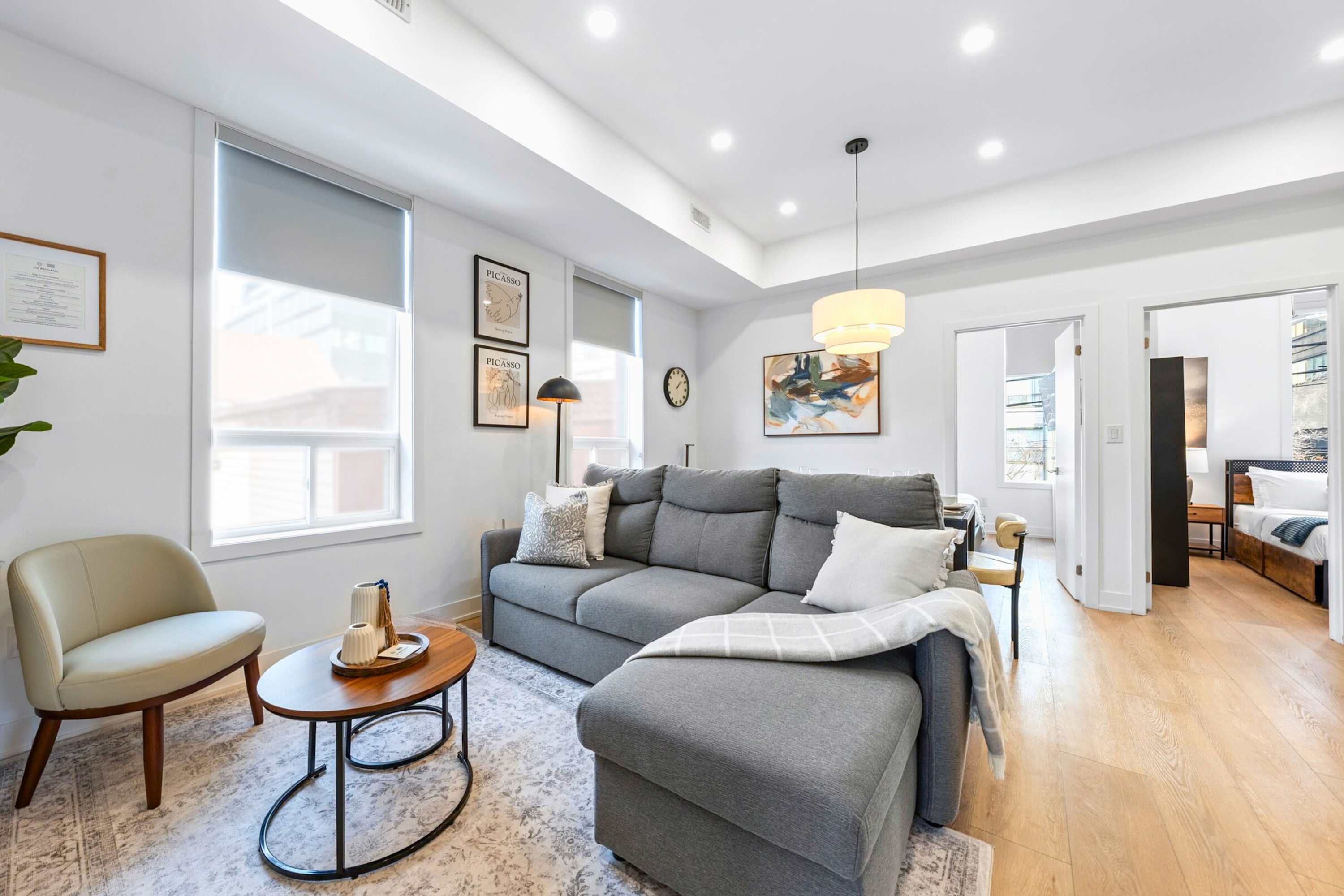 Spacious living room with a grey sectional sofa, a light accent chair, and two round wooden coffee tables on a patterned rug. The room features light wood floors, large windows with roller shades, and framed artwork on the wall.