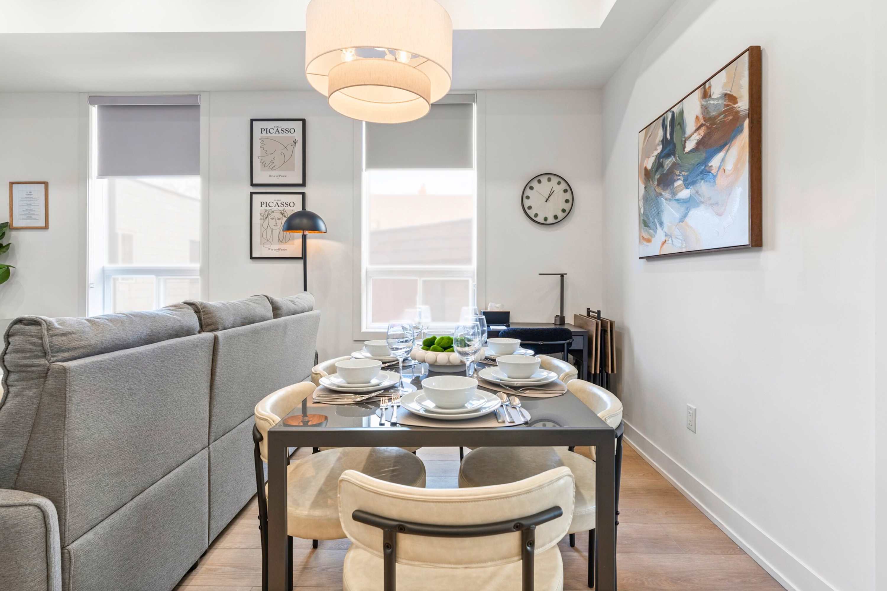 View of the dining area from a slightly different angle, showcasing the dark dining table with four light-colored chairs and a modern drum pendant light. The space is bright with natural light from windows, and a bedroom entrance is visible.
