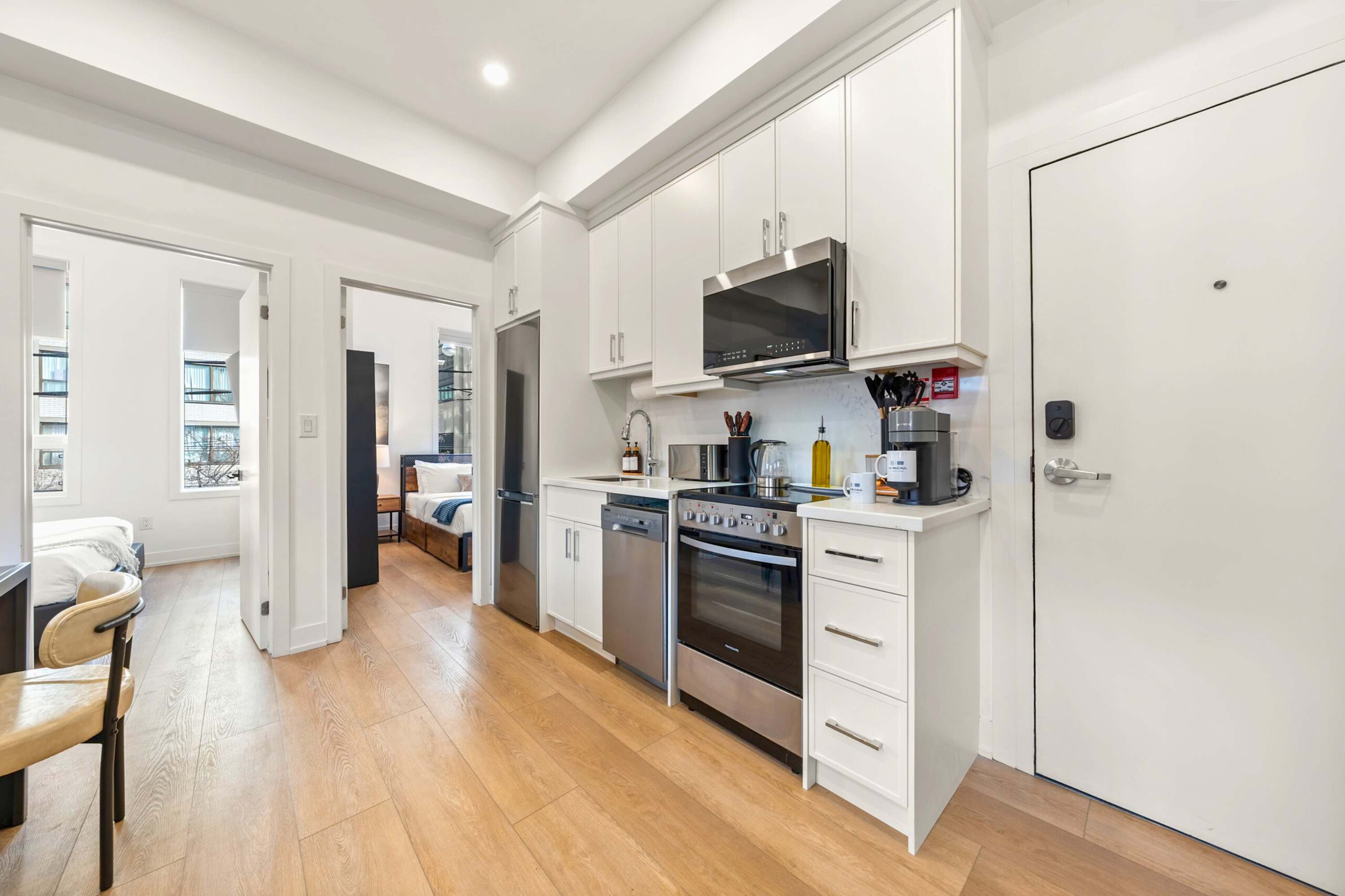 Modern galley kitchen with white shaker-style cabinets, stainless steel appliances including a refrigerator, oven, microwave, and dishwasher. Light hardwood floors extend throughout, and a doorway leads to another room.