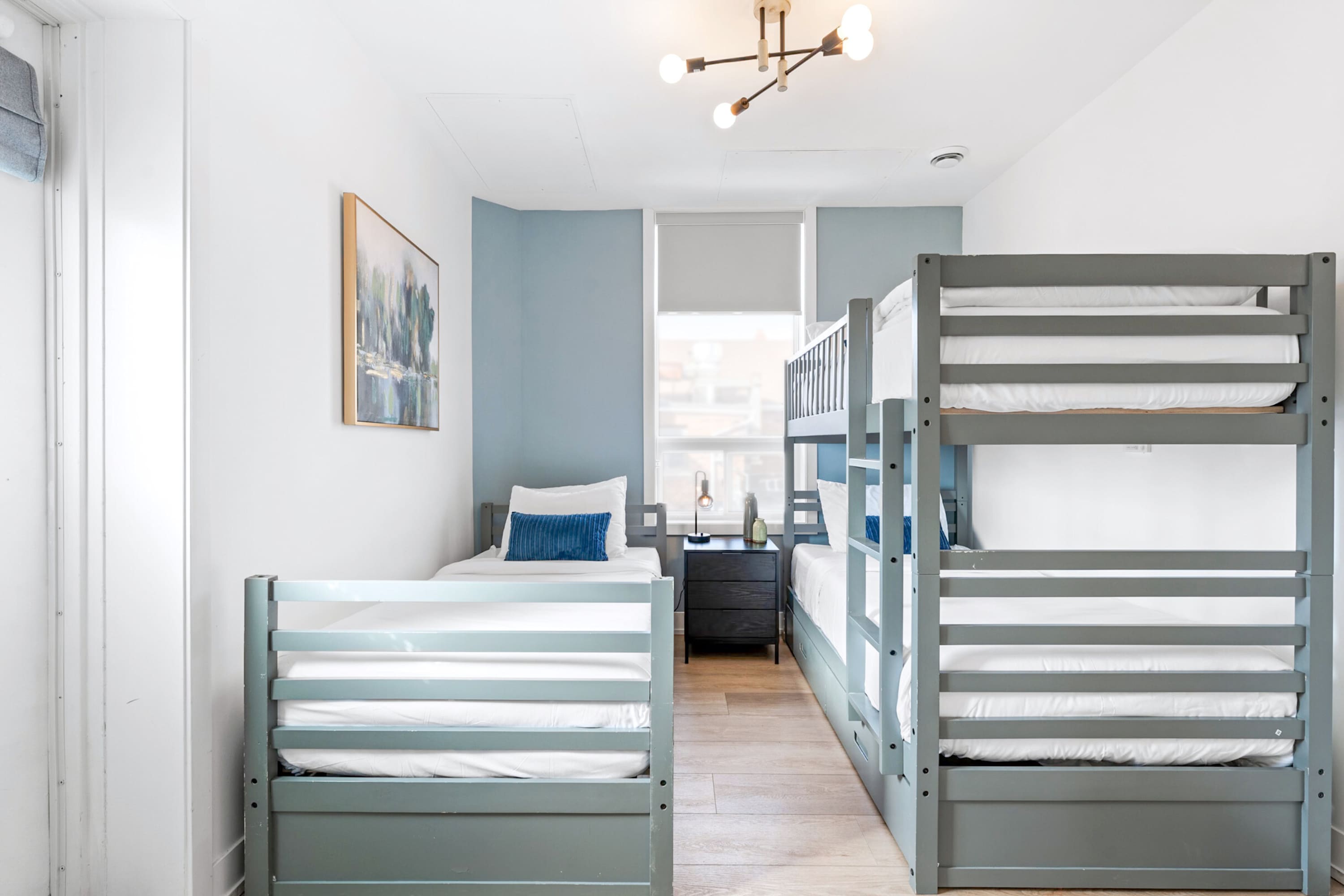 Spacious bedroom with a grey bunk bed and a matching single bed, light hardwood floors, a modern ceiling light, and a window with a blue accent wall.