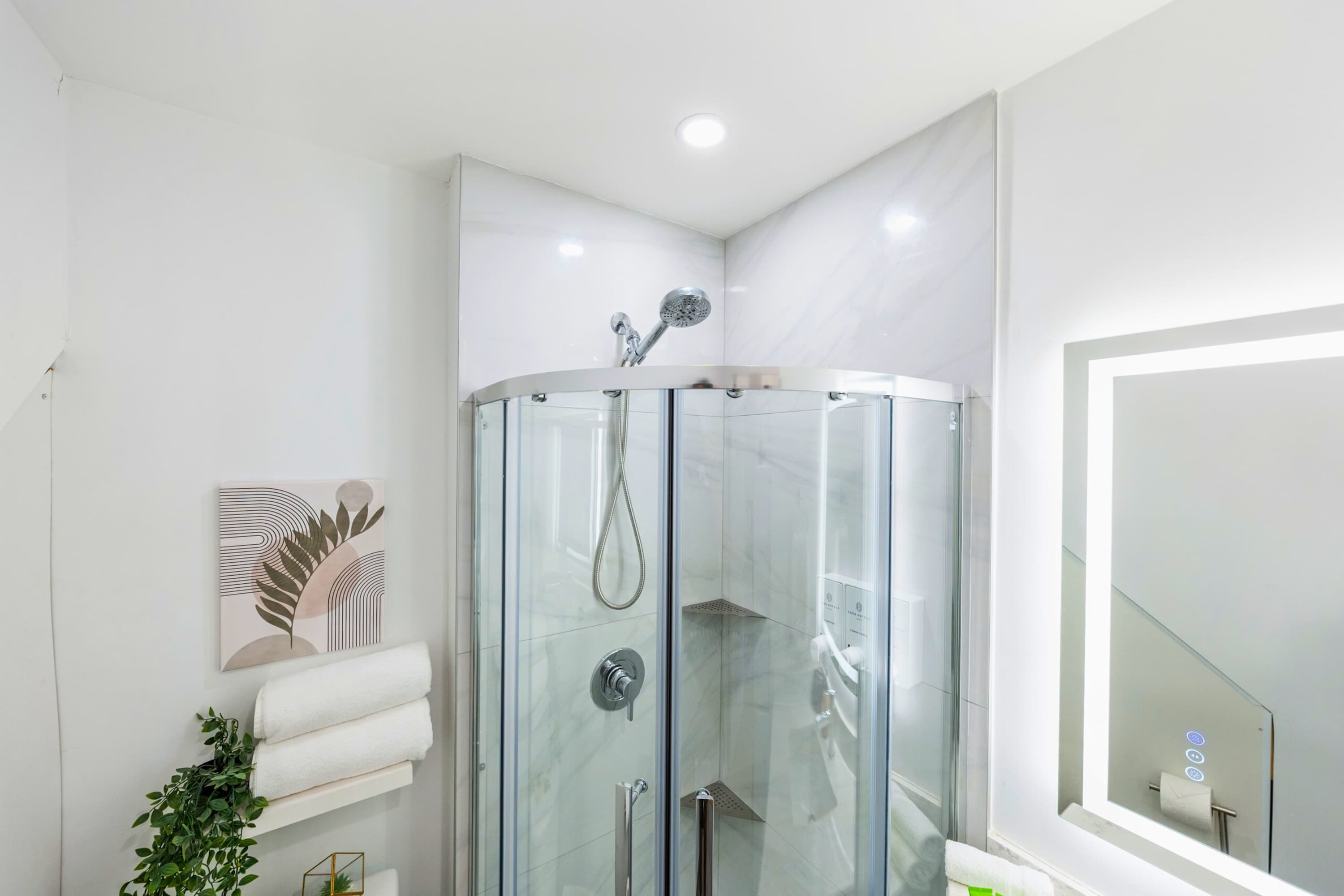 Close-up view of a modern curved glass shower enclosure with a rain shower head and handheld sprayer, set against white marble-look tiled walls.