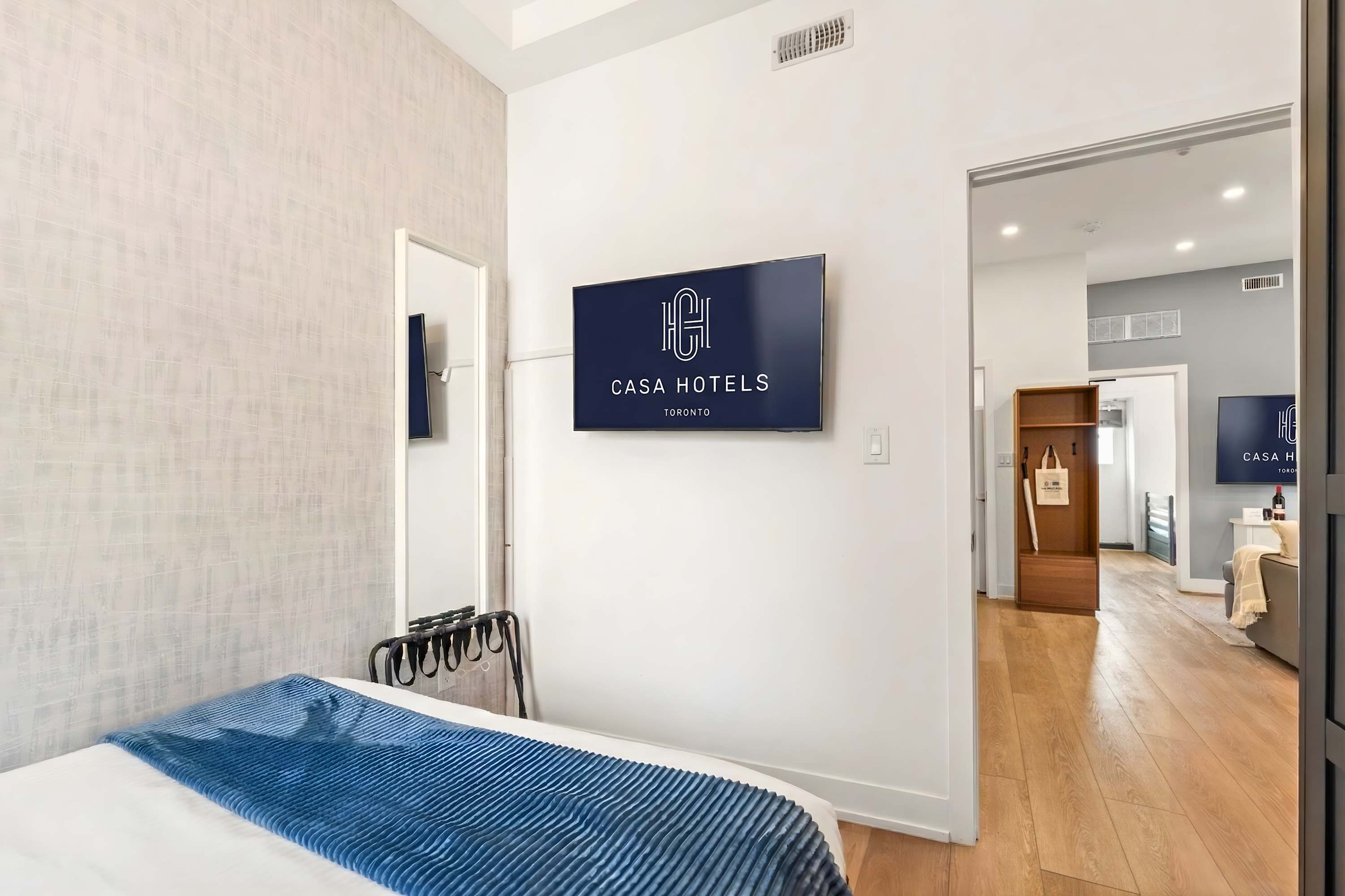 Bedroom wall with a mounted flat-screen TV displaying "CASA HOTELS TORONTO" logo, a full-length mirror, and a luggage rack, with hardwood floors.