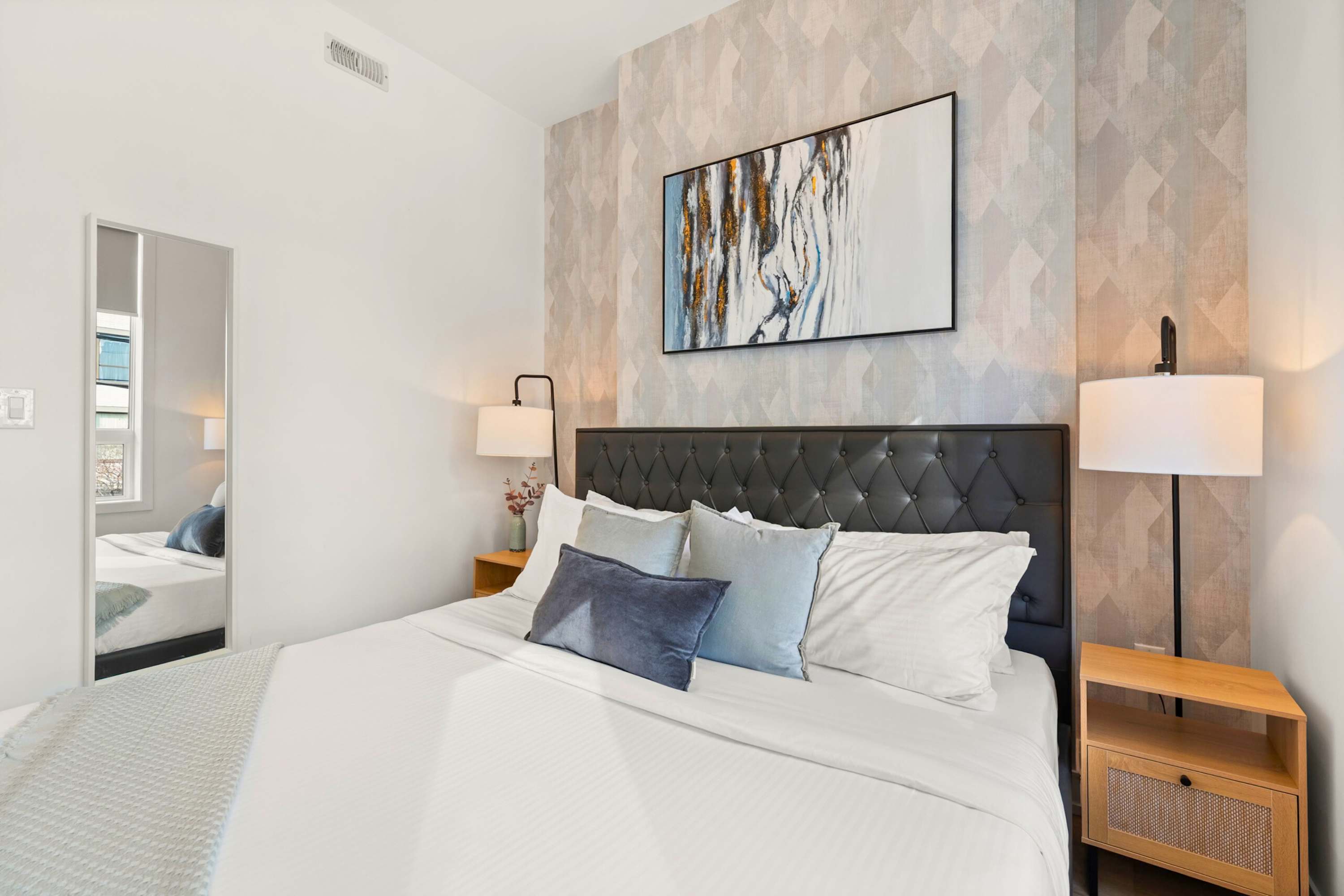 Cozy bedroom with a queen bed, a black tufted headboard, and light wooden nightstands with lamps. The room features a patterned accent wall behind the bed and a full-length mirror reflecting the opposite side of the room.