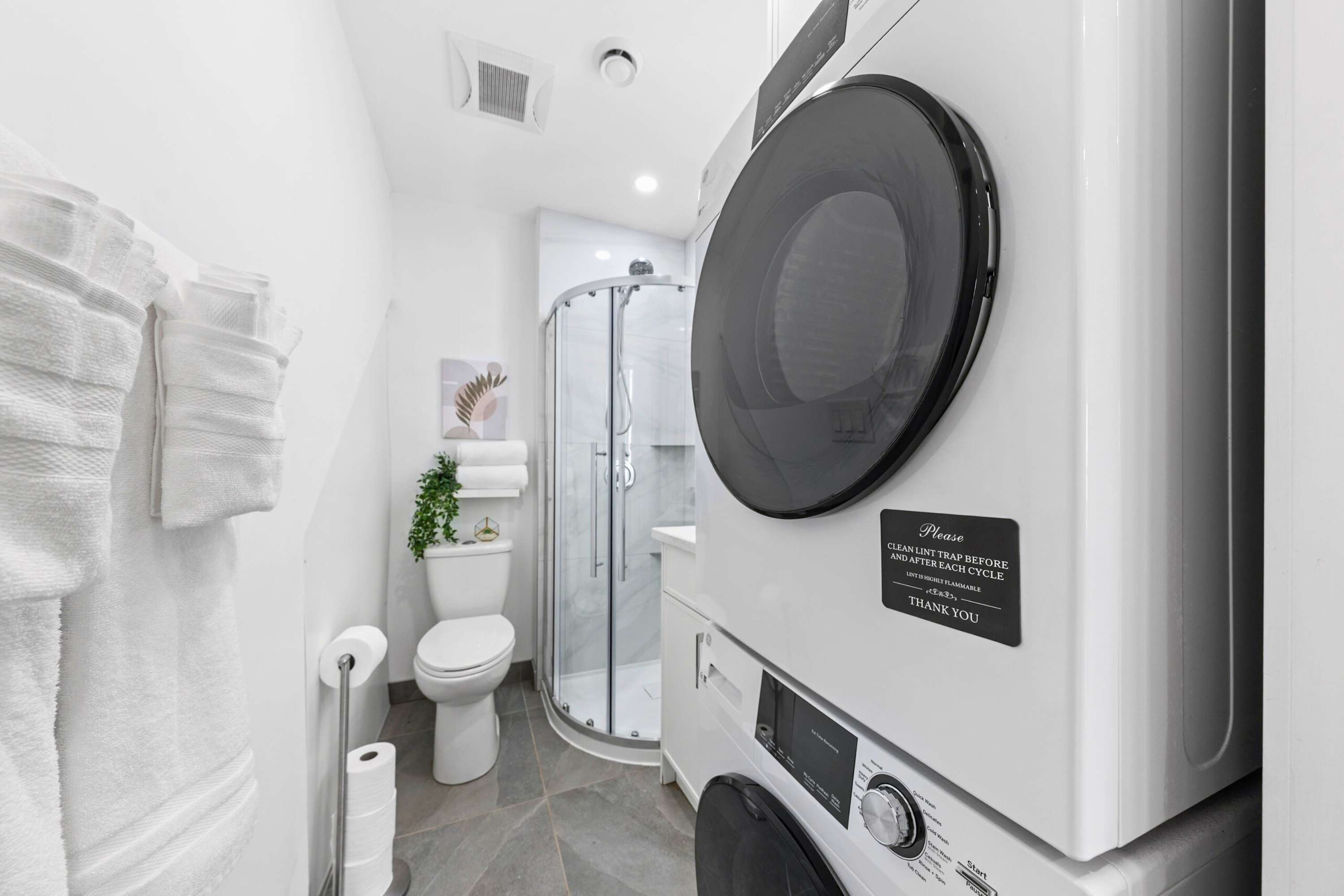 Compact laundry area with a stacked white washer and dryer unit, located in a hallway or alcove with dark tiled floors, adjacent to a bathroom.