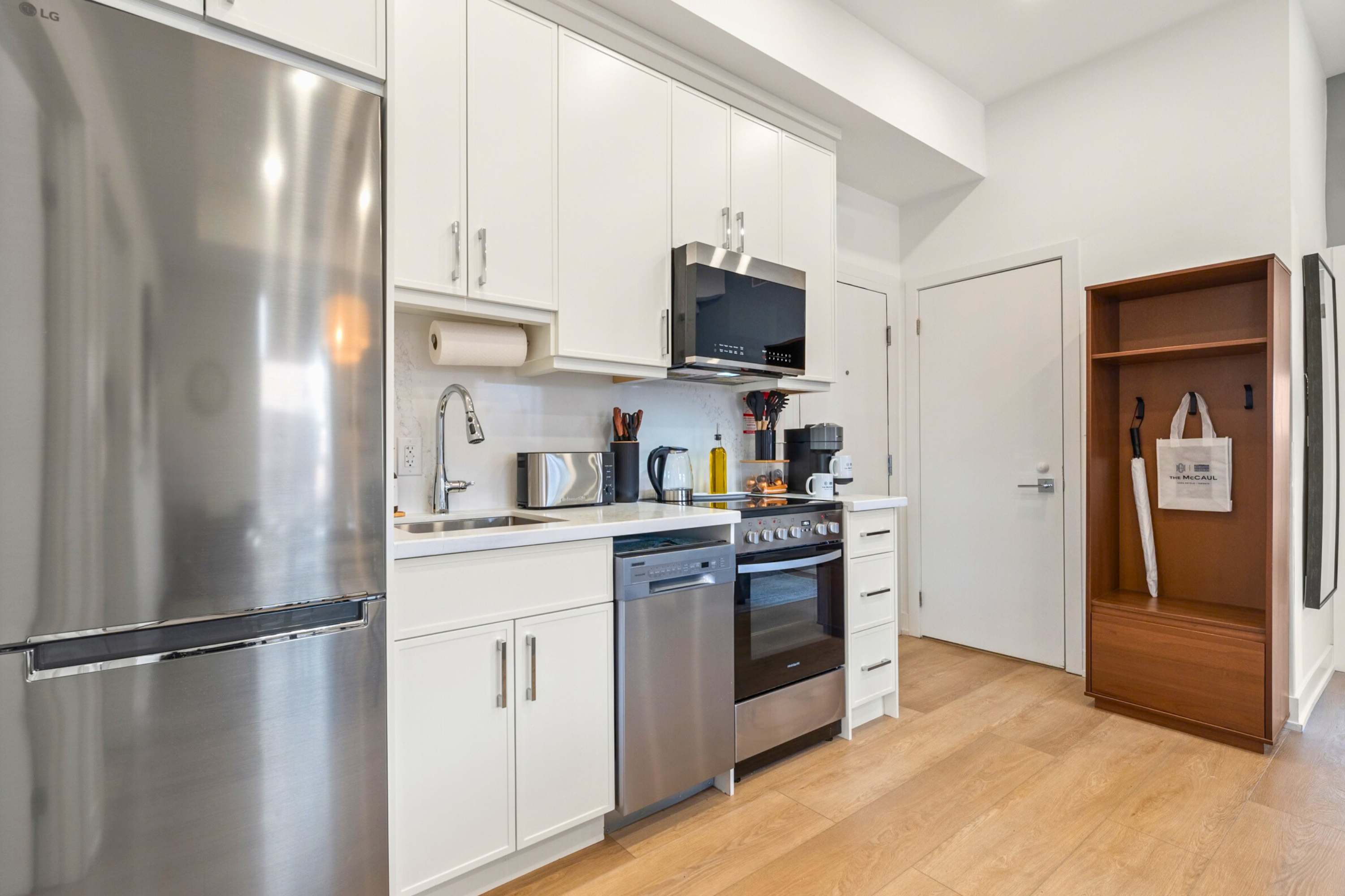 Well-equipped kitchen featuring white cabinetry, stainless steel appliances, and light hardwood flooring. A coffee maker, toaster, and other kitchen essentials are visible on the white countertop, with a small entryway bench to the right.