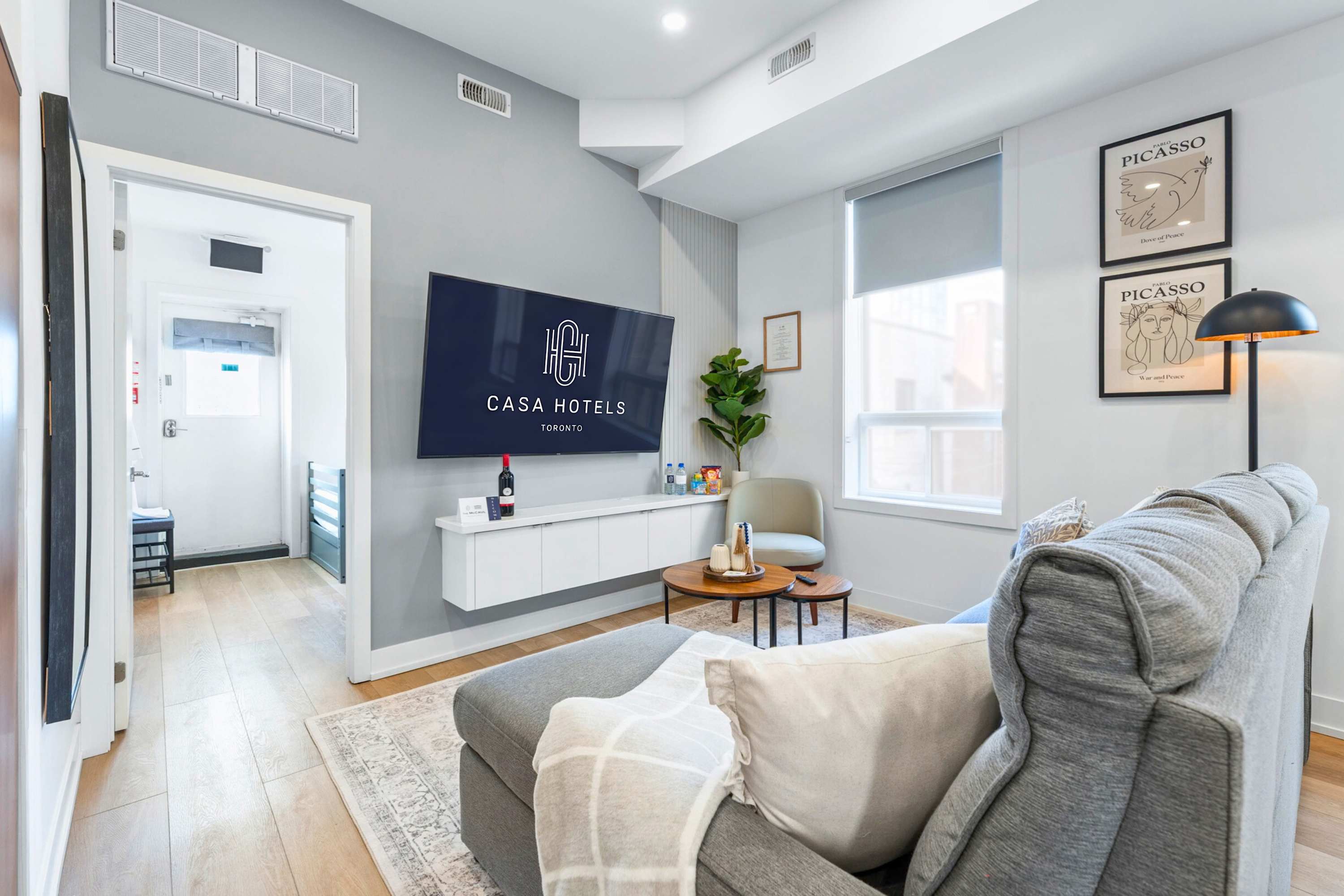 View of the living room from the sectional sofa, showing a wall-mounted TV with "CASA HOTELS" branding above a white floating console. A light accent chair, nesting coffee tables, and an indoor plant are visible, with a doorway leading to another room.