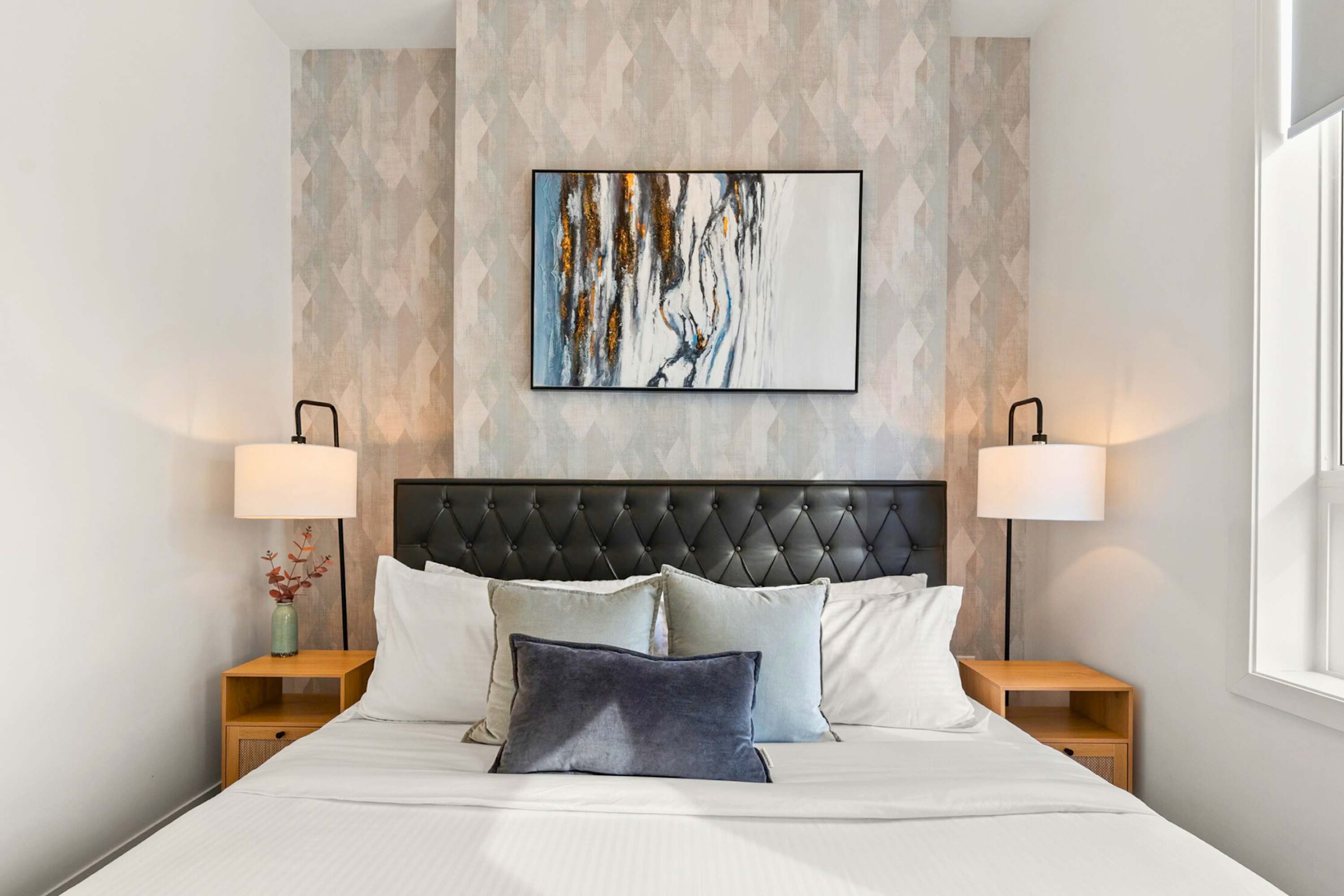 Serene bedroom featuring a queen-sized bed with a tufted black headboard, flanked by wooden nightstands with modern lamps. A large abstract painting hangs above the bed, complemented by a patterned accent wall.