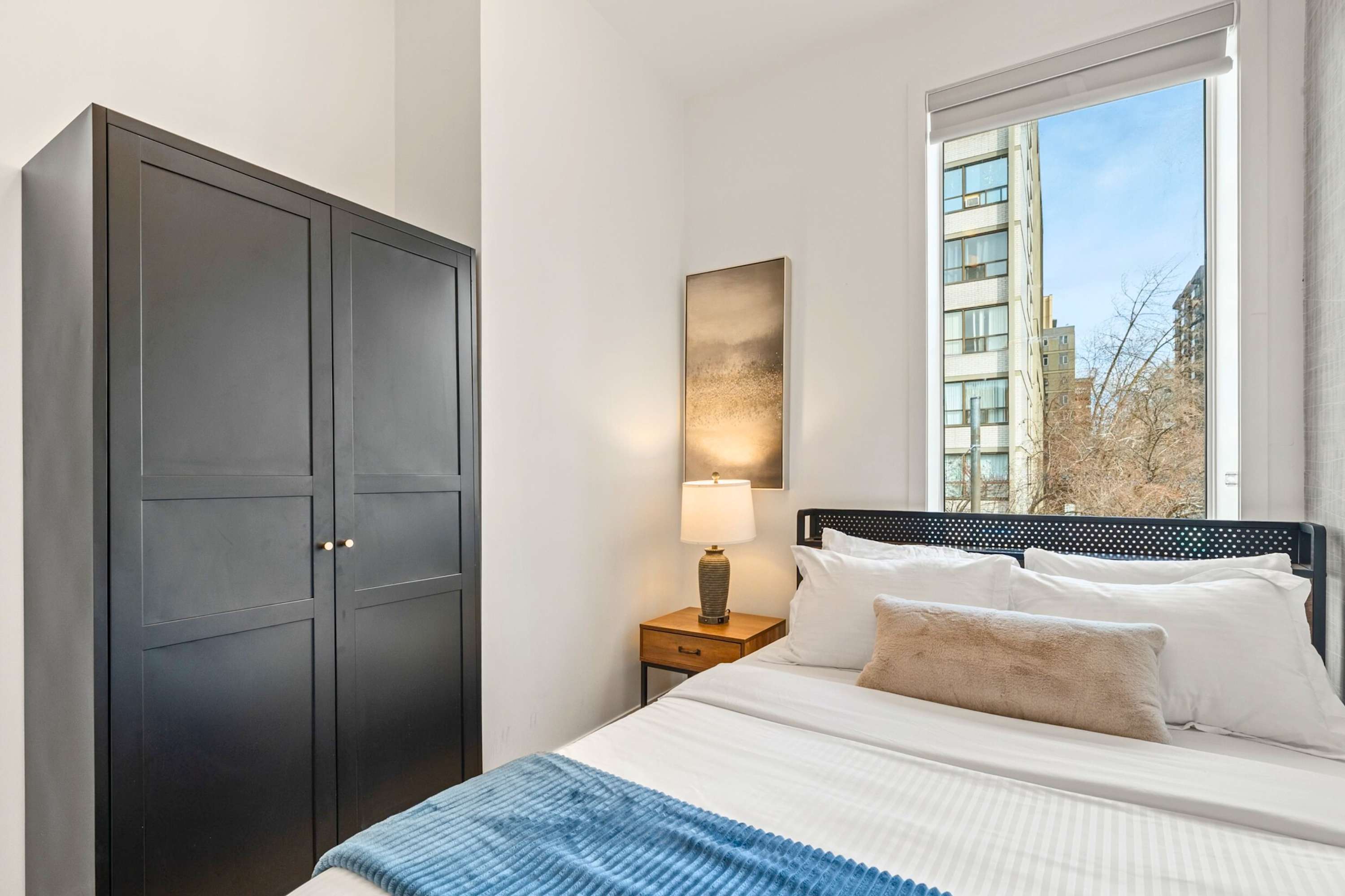 View of a bedroom with a queen bed, a dark wooden wardrobe, a wooden nightstand with a lamp, and a large window overlooking city buildings.
