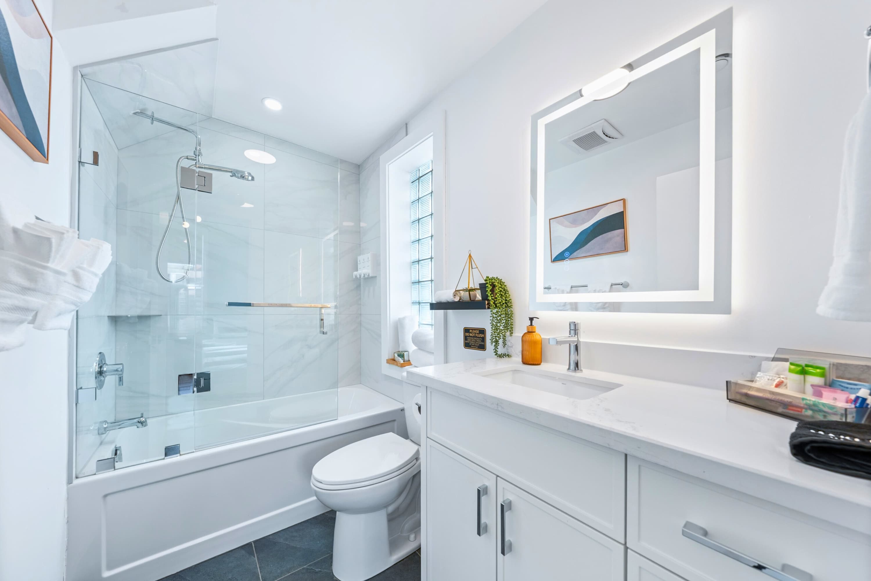 A modern white bathroom features a bathtub with a glass shower enclosure, a white vanity with a single sink, and an illuminated mirror. A small window with glass blocks provides natural light.