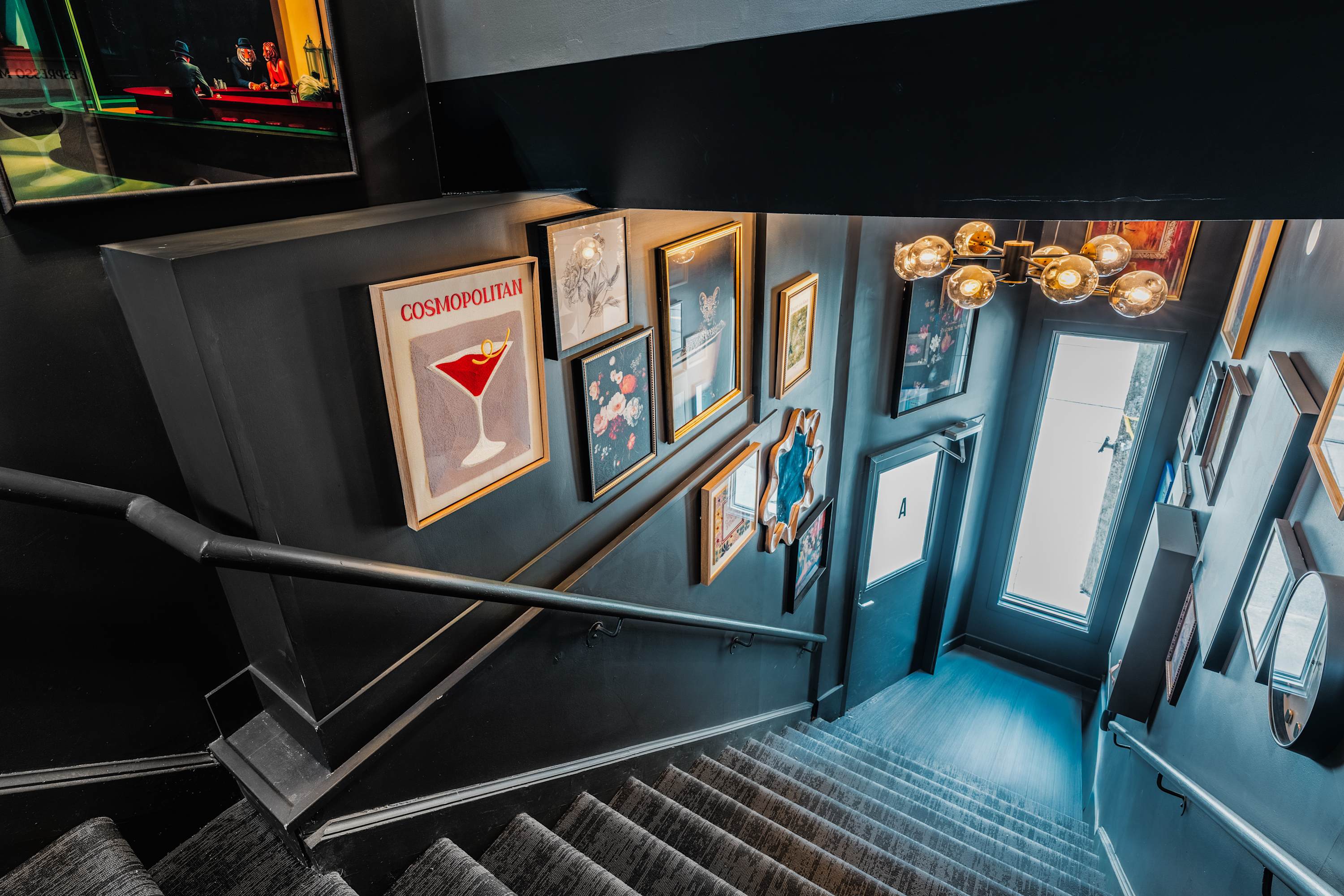 A top-down view of a dark-walled staircase with a carpet runner, showcasing a gallery wall of diverse framed art and mirrors. A large window at the bottom of the stairs provides natural light.