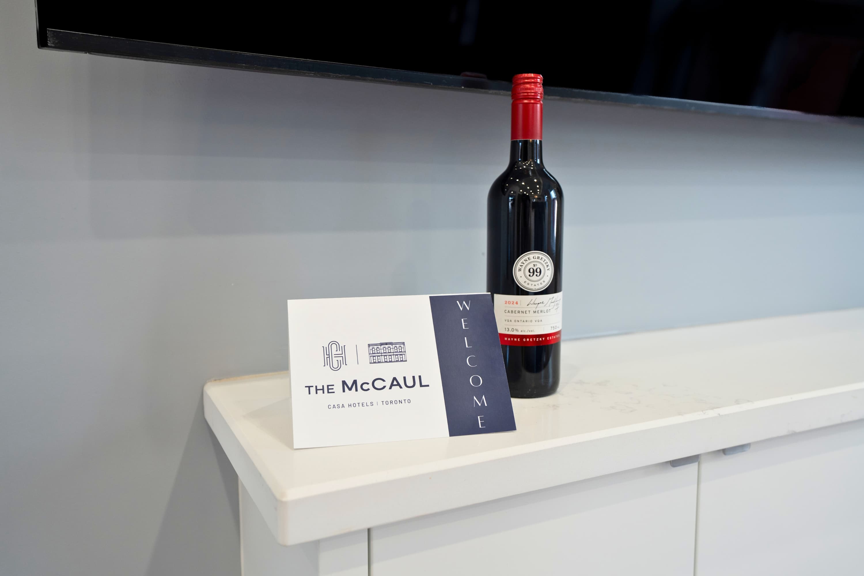 Detail shot of a welcome gift on a white floating console, featuring a bottle of red wine and a "WELCOME THE MCCAUL By Casa Hotels" card. The background shows a wall-mounted TV with the "CASA HOTELS" logo.