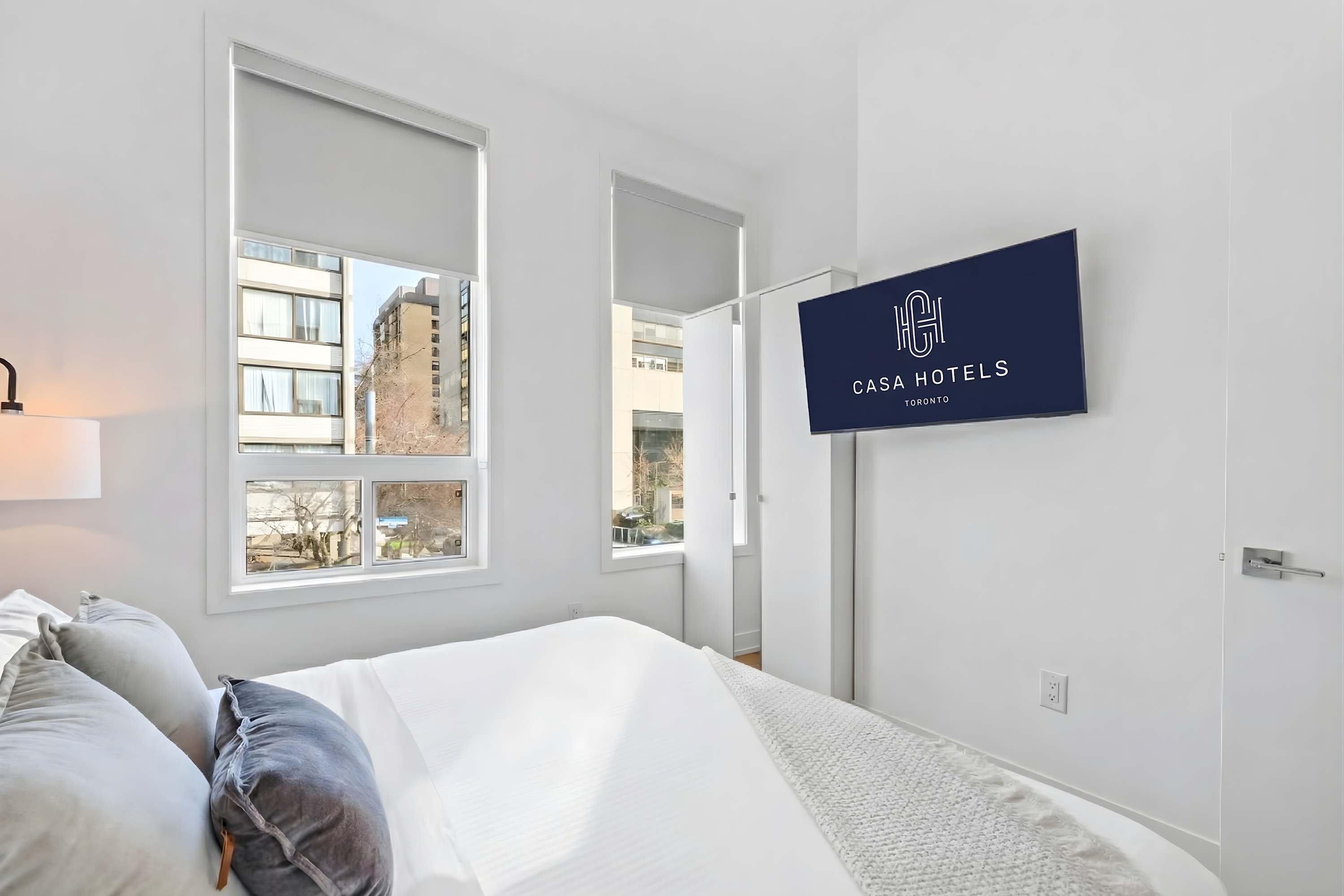 Bright bedroom with a queen bed dressed in white linens and blue accent pillows, featuring large windows with roller shades. A wall-mounted TV displays "CASA HOTELS", and light hardwood floors complete the modern aesthetic.