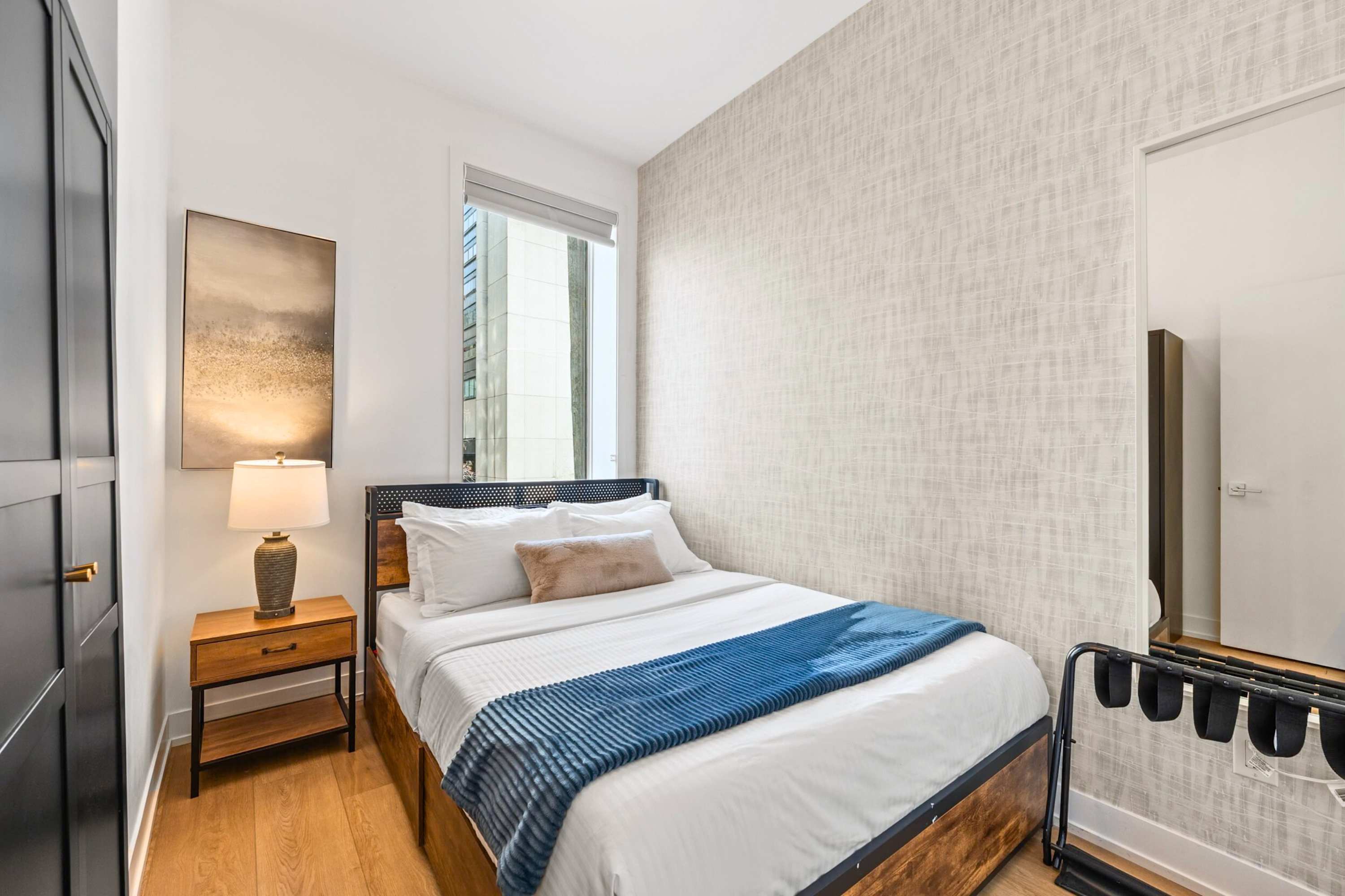 Cozy bedroom with a queen-sized bed featuring a dark metal headboard, a wooden nightstand with a lamp, and a large window offering a city view.